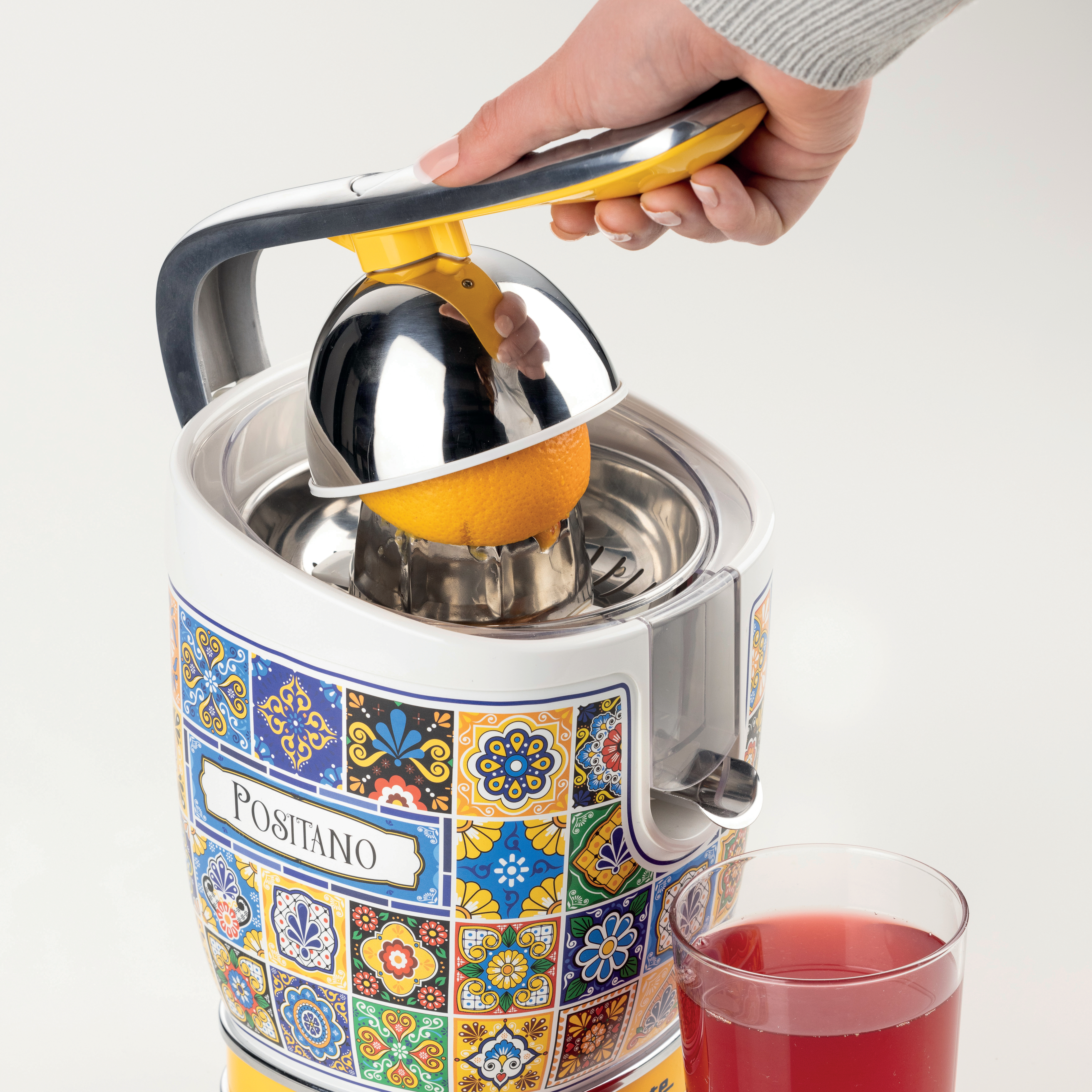 Eine Hand presst eine Orange mit einer Saftpresse aus. Neben der Saftpresse steht ein Glas Saft. Die Saftpresse hat ein farbenfrohes Muster.