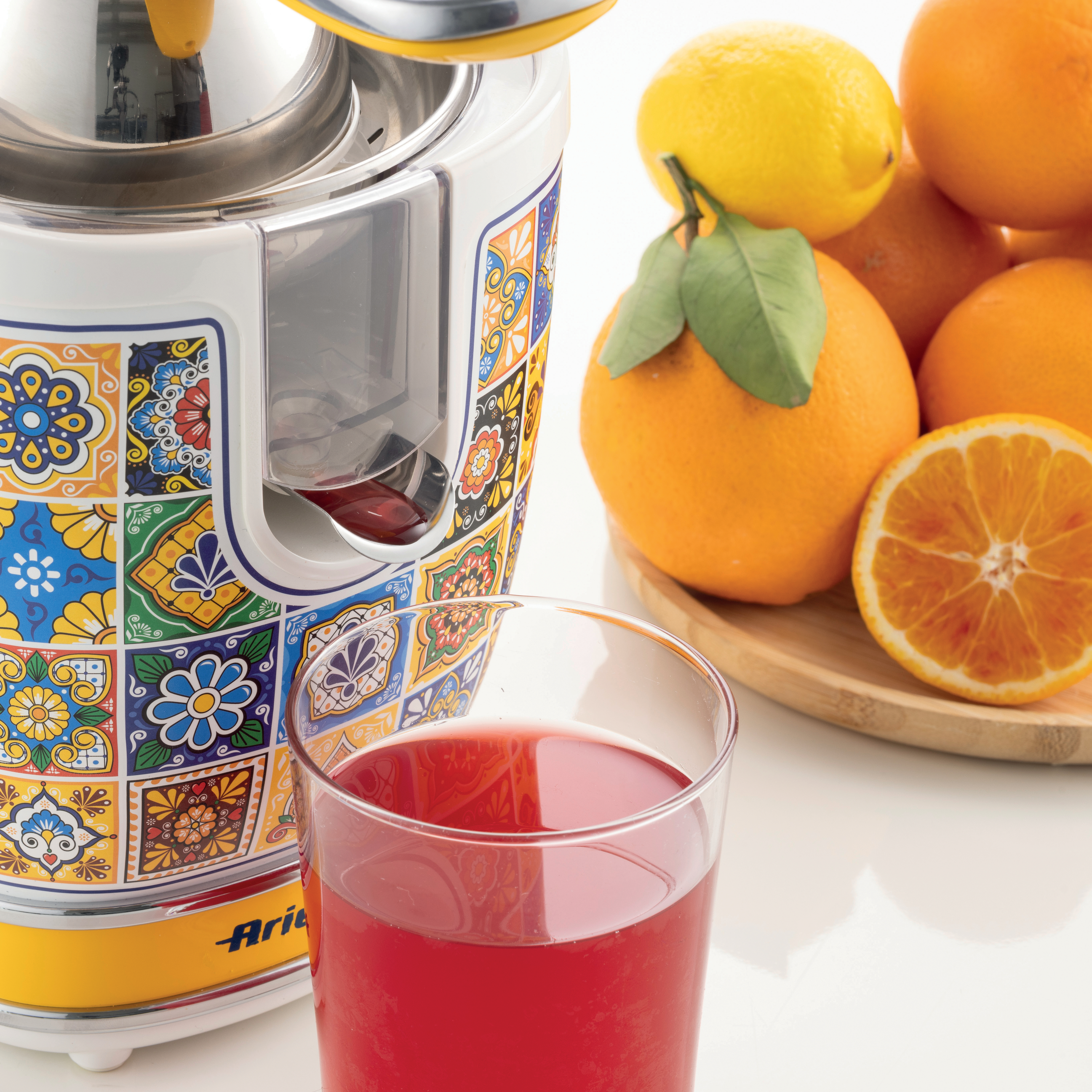 Ein Entsafter mit bunten Fliesen steht neben einem Glas Saft. Orangen, Zitronen und eine halbe Orange sind in der Nähe.