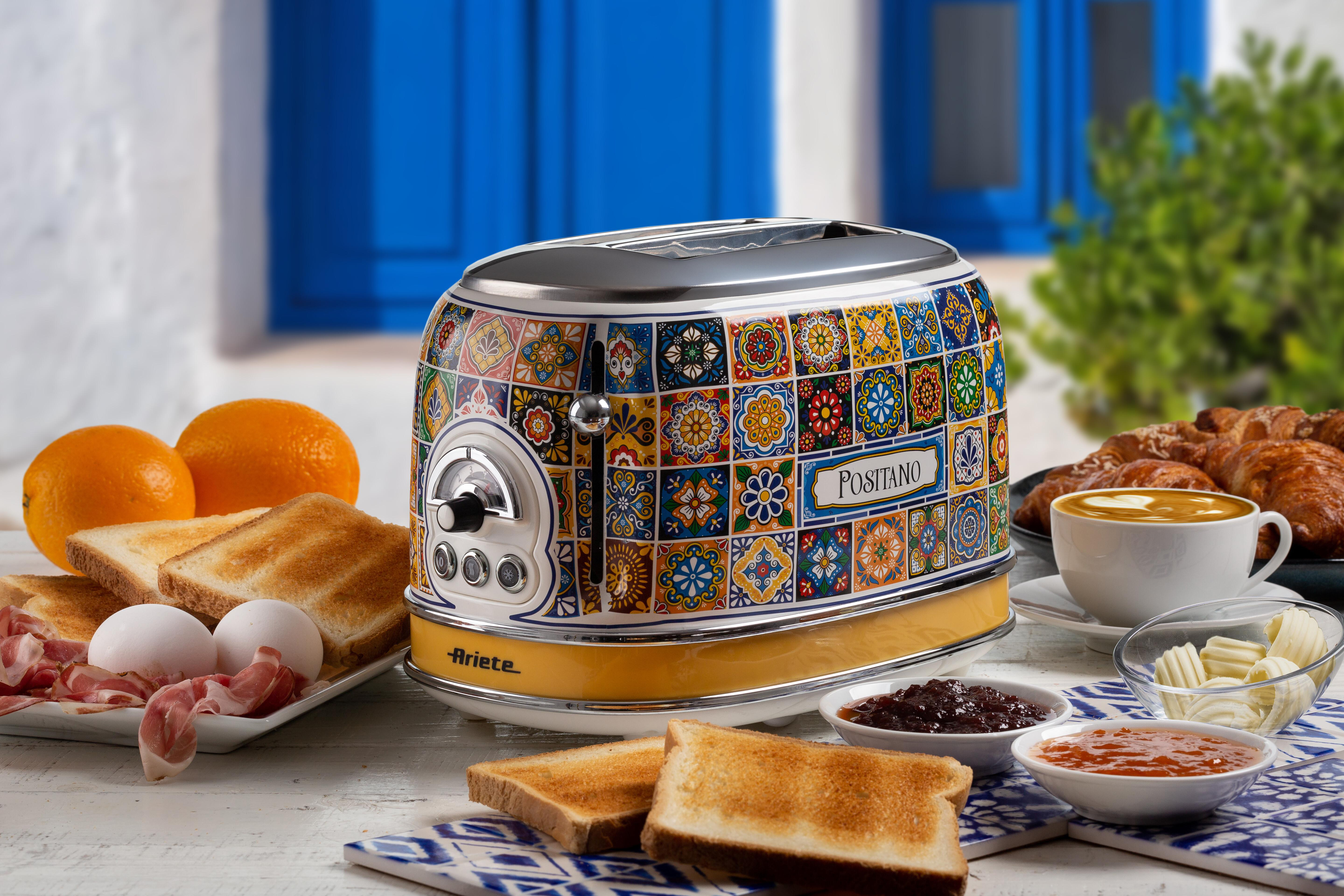Auf einem Tisch stehen ein Toaster, Frühstücksessen und Kaffee. Orangen, Eier, Speck, Toast und Marmelade sind sichtbar.