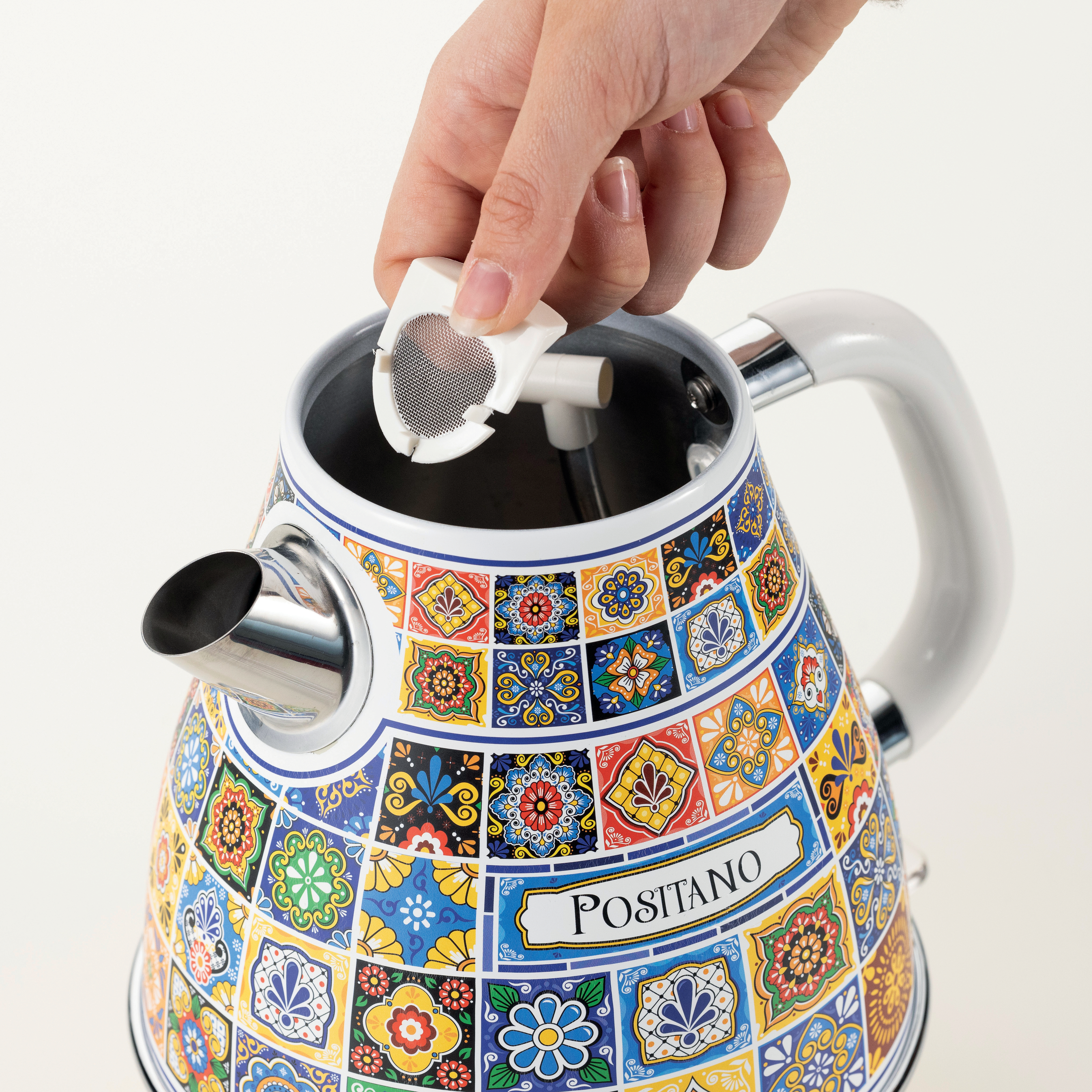 Eine Hand, die einen Tee-Infuser in einen bunten Wasserkocher legt. Der Wasserkocher hat ein florales Fliesendesign mit dem Wort 'Positano'.