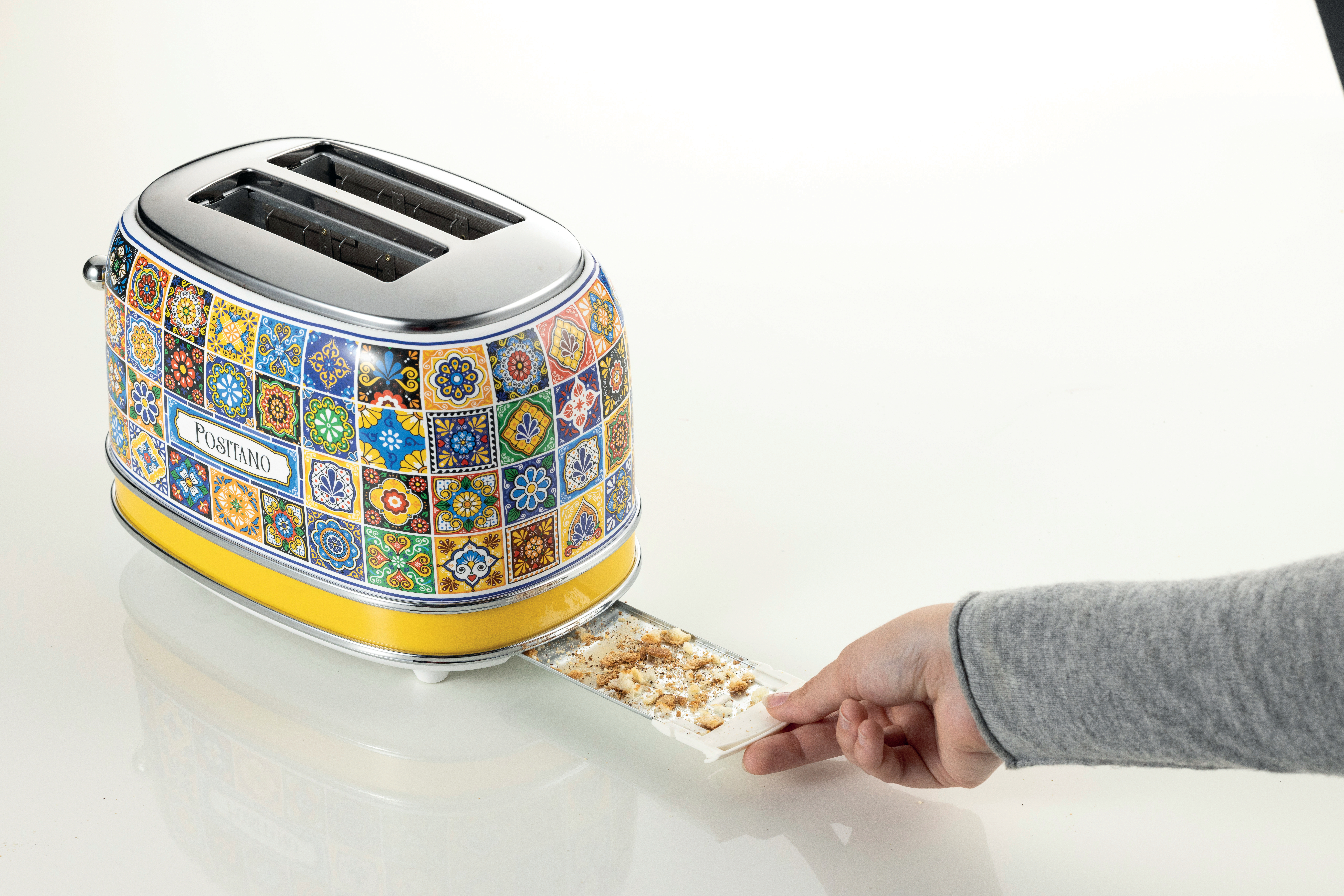 Ein bunter Toaster mit einer Hand, die das Krümelblech herausnimmt. Es steht auf einer weißen Oberfläche. Der Toaster hat ein 'Positano'-Etikett.