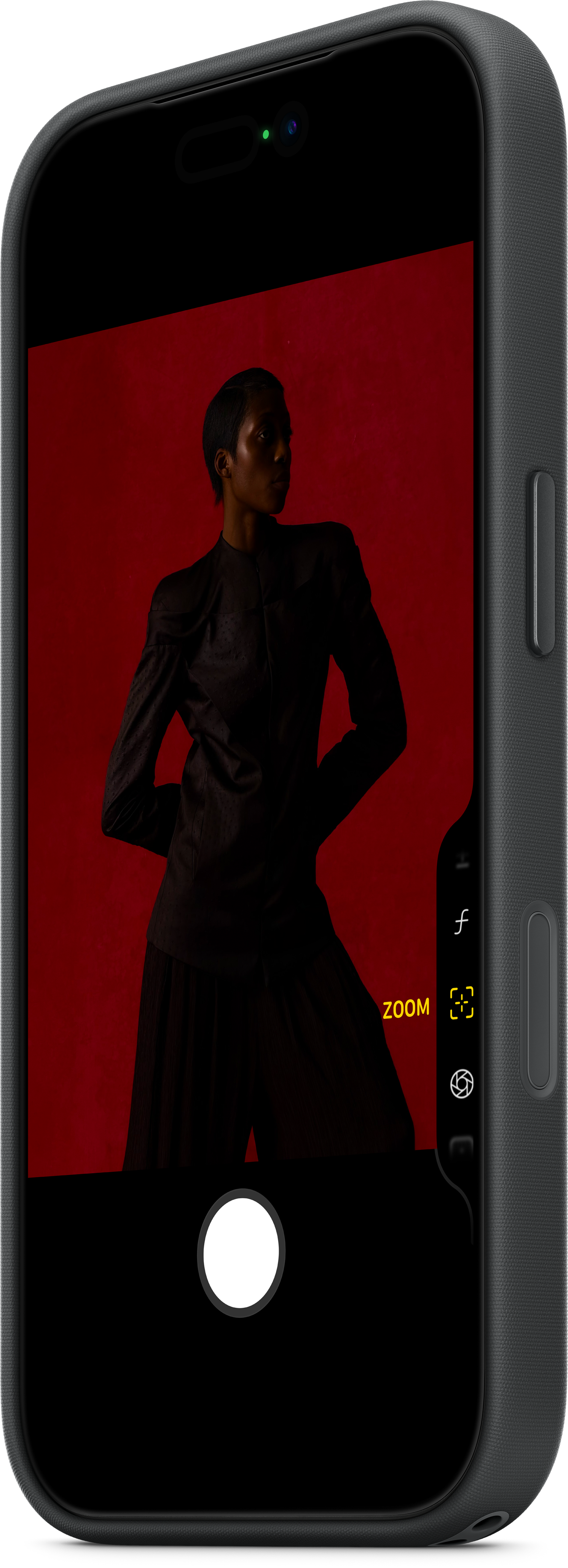 Una persona in abiti scuri posa davanti a uno sfondo rosso. L'immagine è incorniciata da un telefono che mostra un'interfaccia fotocamera.