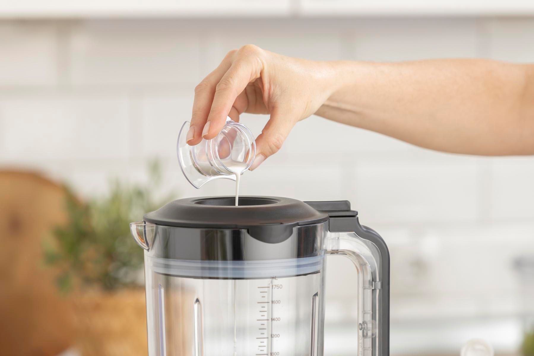 Eine Hand gießt eine weiße Flüssigkeit aus einem kleinen Messbecher in einen Mixer. Der Mixer ist schwarz und transparent. Der Hintergrund ist weiß.