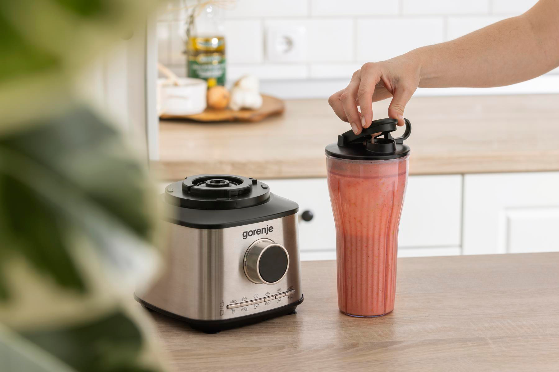 Eine Person öffnet einen Mixerbecher mit rosa Smoothie. Ein Gorenje-Mixer steht auf einer Holztheke, Küche im Hintergrund.