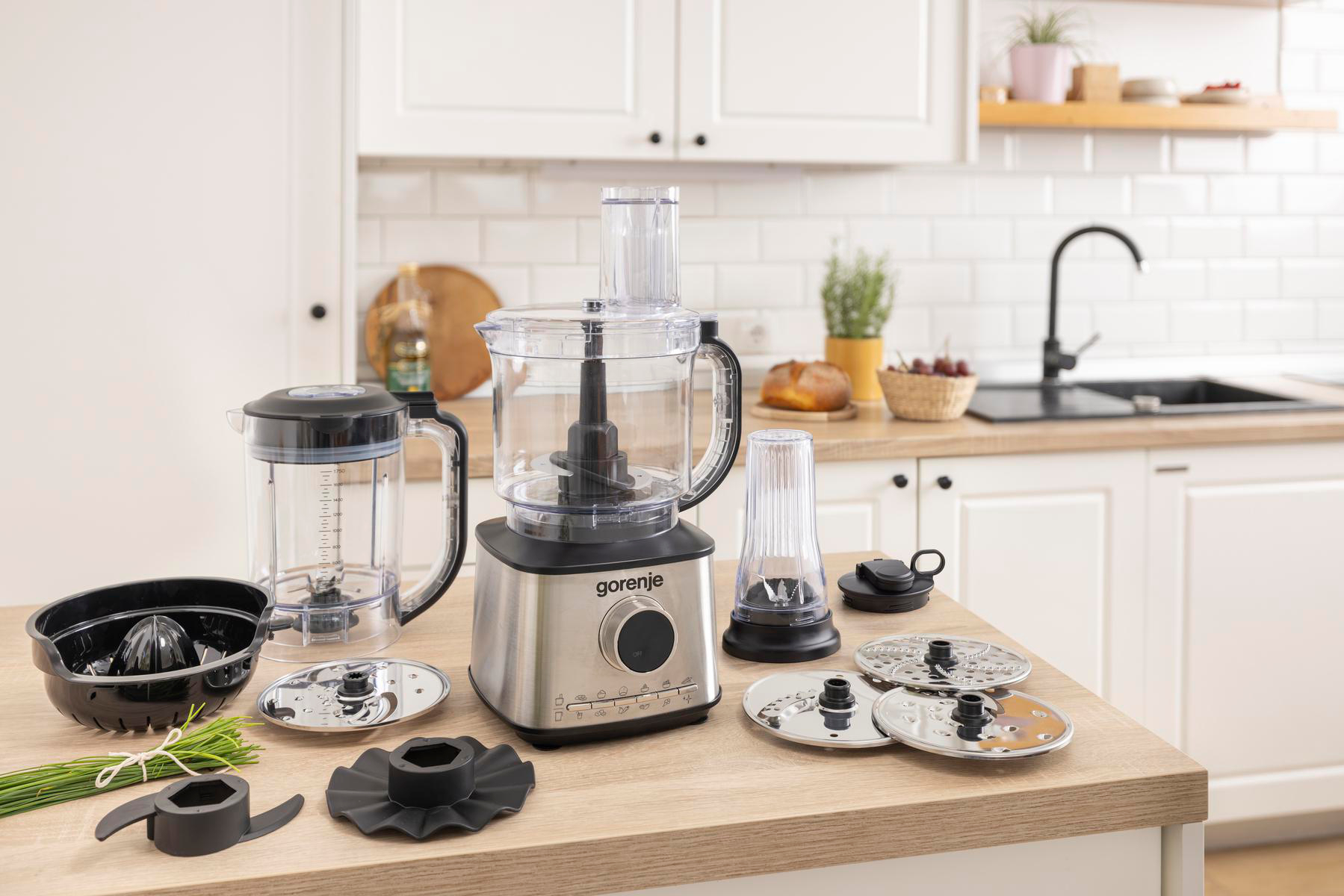 Eine Küchenarbeitsplatte mit einem Foodprocessor, Mixer und verschiedenen Aufsätzen. Der Hintergrund umfasst Küchenelemente wie Spüle und Brot.