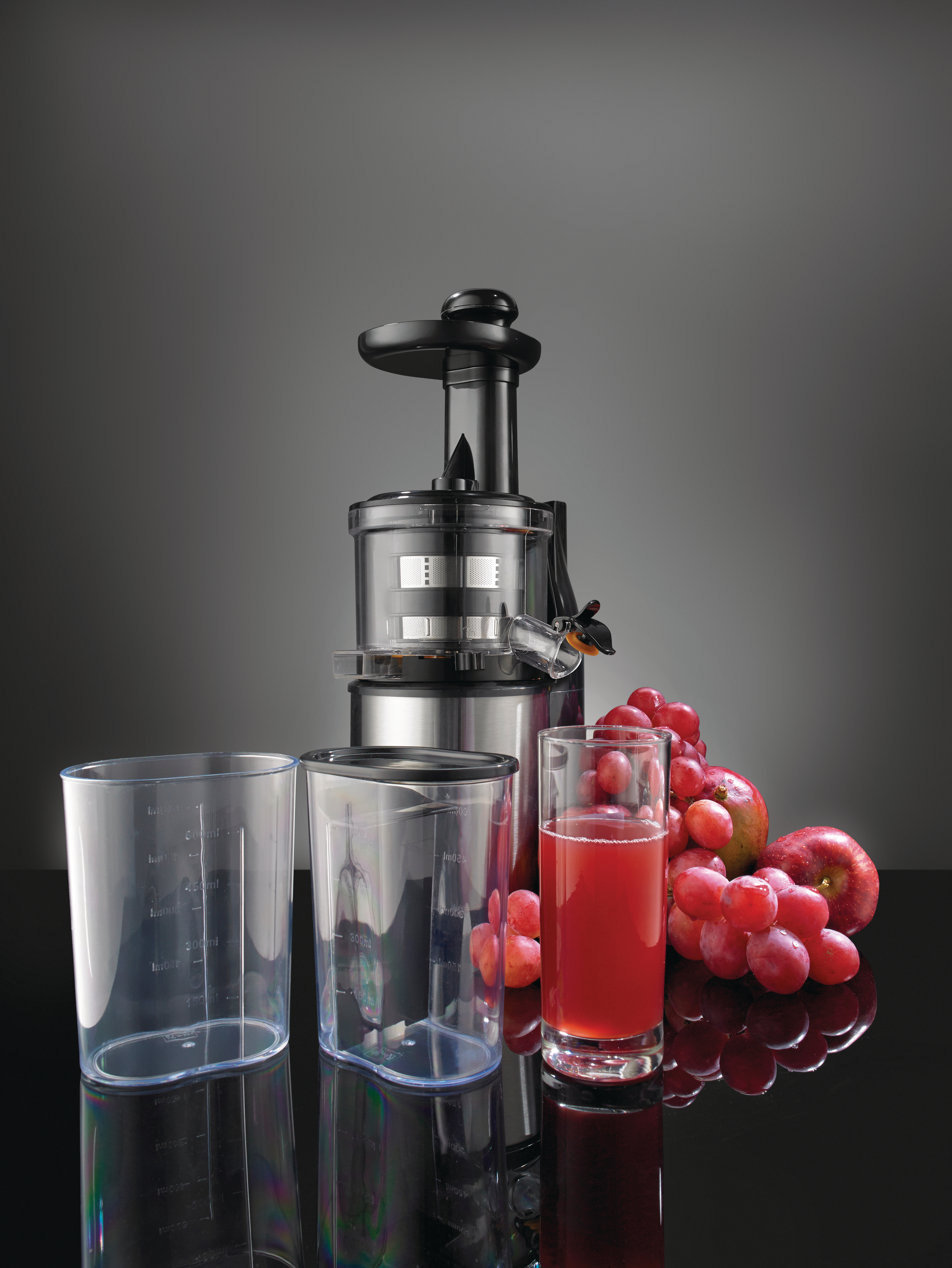 Ein Entsafter mit zwei durchsichtigen Behältern, einem Glas Saft, Trauben und Äpfeln. Der Hintergrund ist grau.