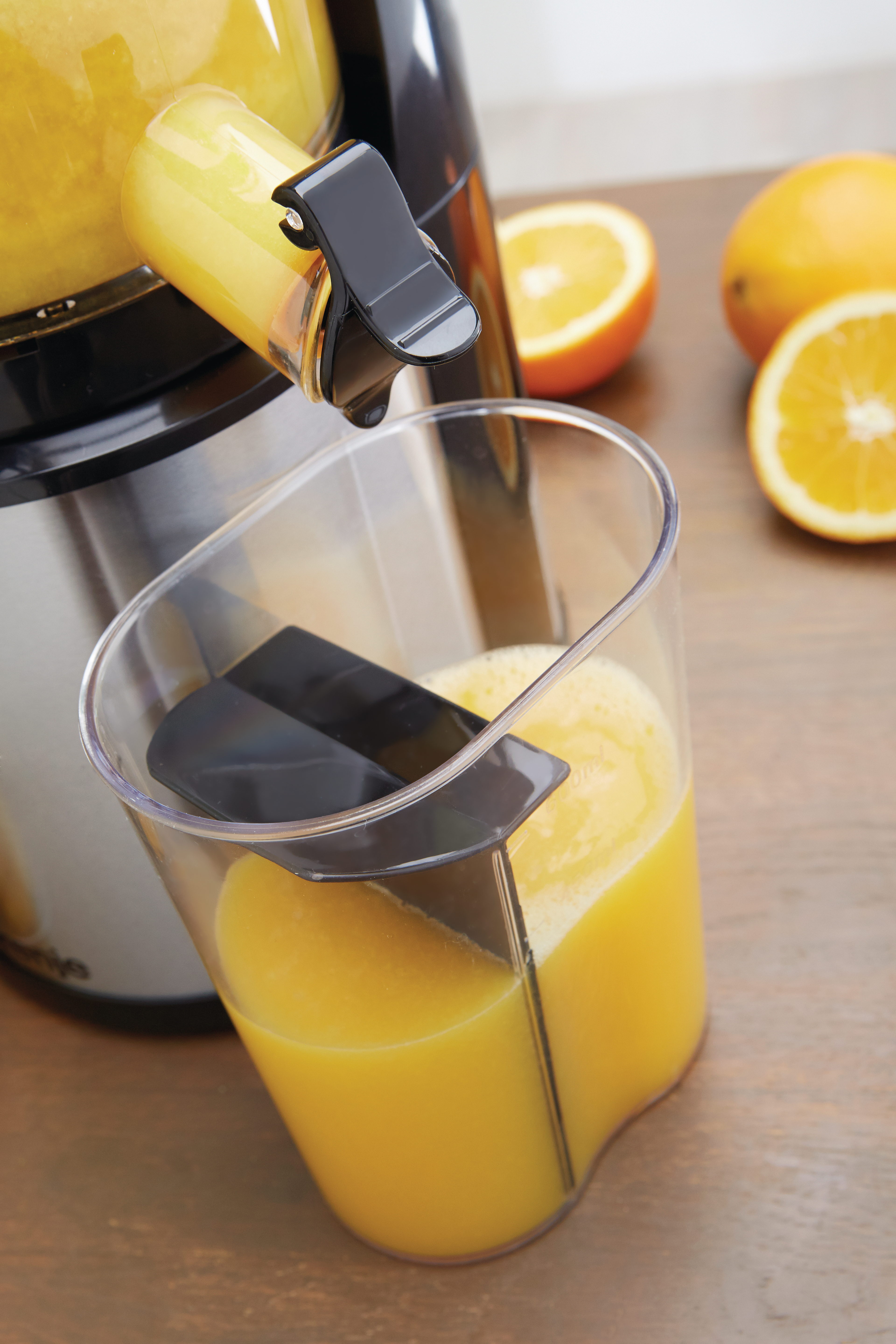 Ein Entsafter, der frischen Orangensaft in ein klares Glas abgibt. Orangen liegen auf dem Holztisch. Der Entsafter ist schwarz und aus Edelstahl.