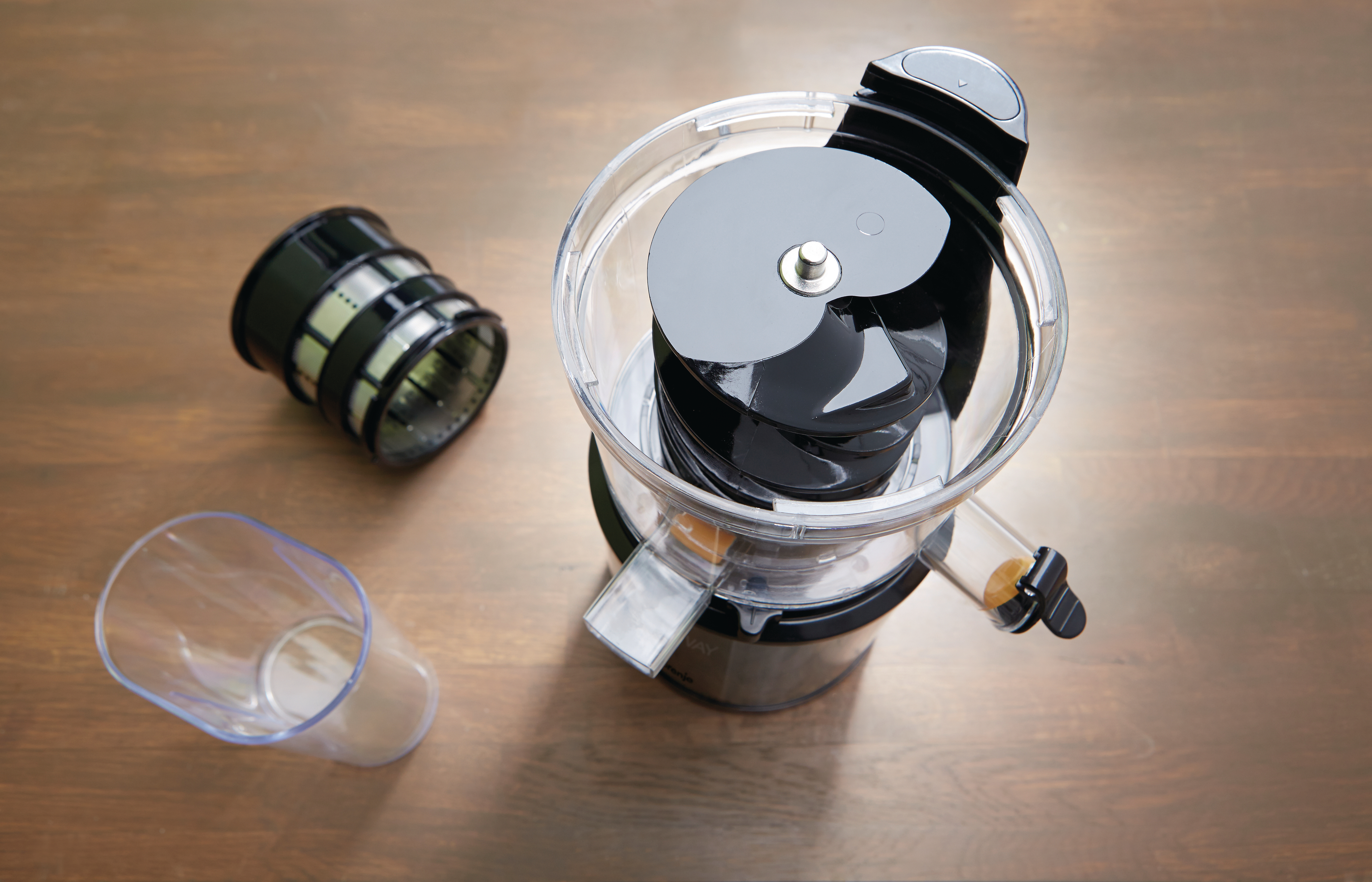 Teile einer Saftpresse auf einer Holzoberfläche. Enthält eine zerlegte Saftpresse, einen Filter und einen durchsichtigen Plastikbecher. Die Maschine ist schwarz und transparent.