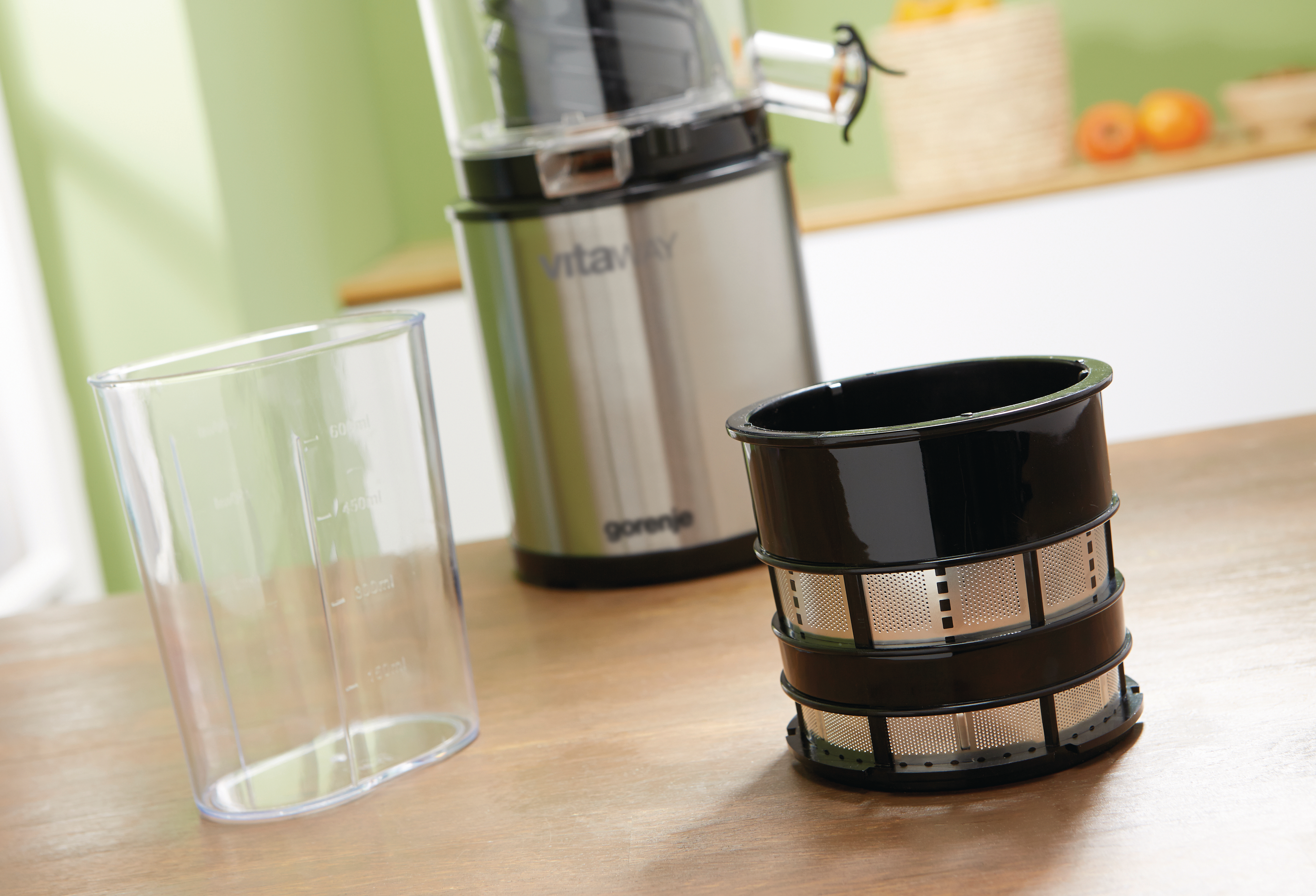 Eine Saftpresse mit Teilen auf einem Holztisch. Ein klarer Becher, ein schwarzer Filter und die Saftpresse. Der Hintergrund hat Obst und eine grüne Wand.