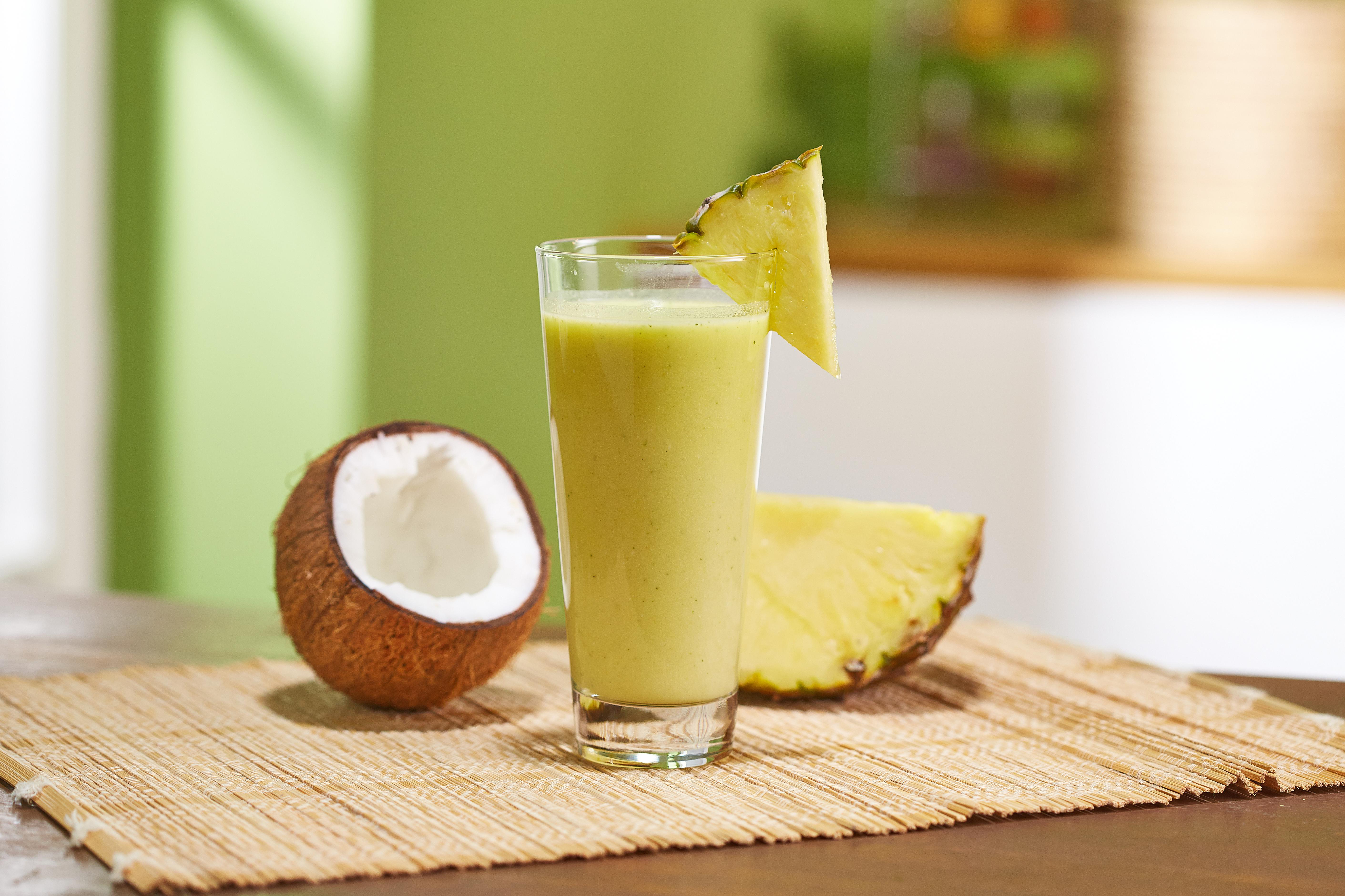 Ein Glas gelber Smoothie mit Ananas-Keil, Kokosnuss und Ananasscheibe auf einer braunen Matte.