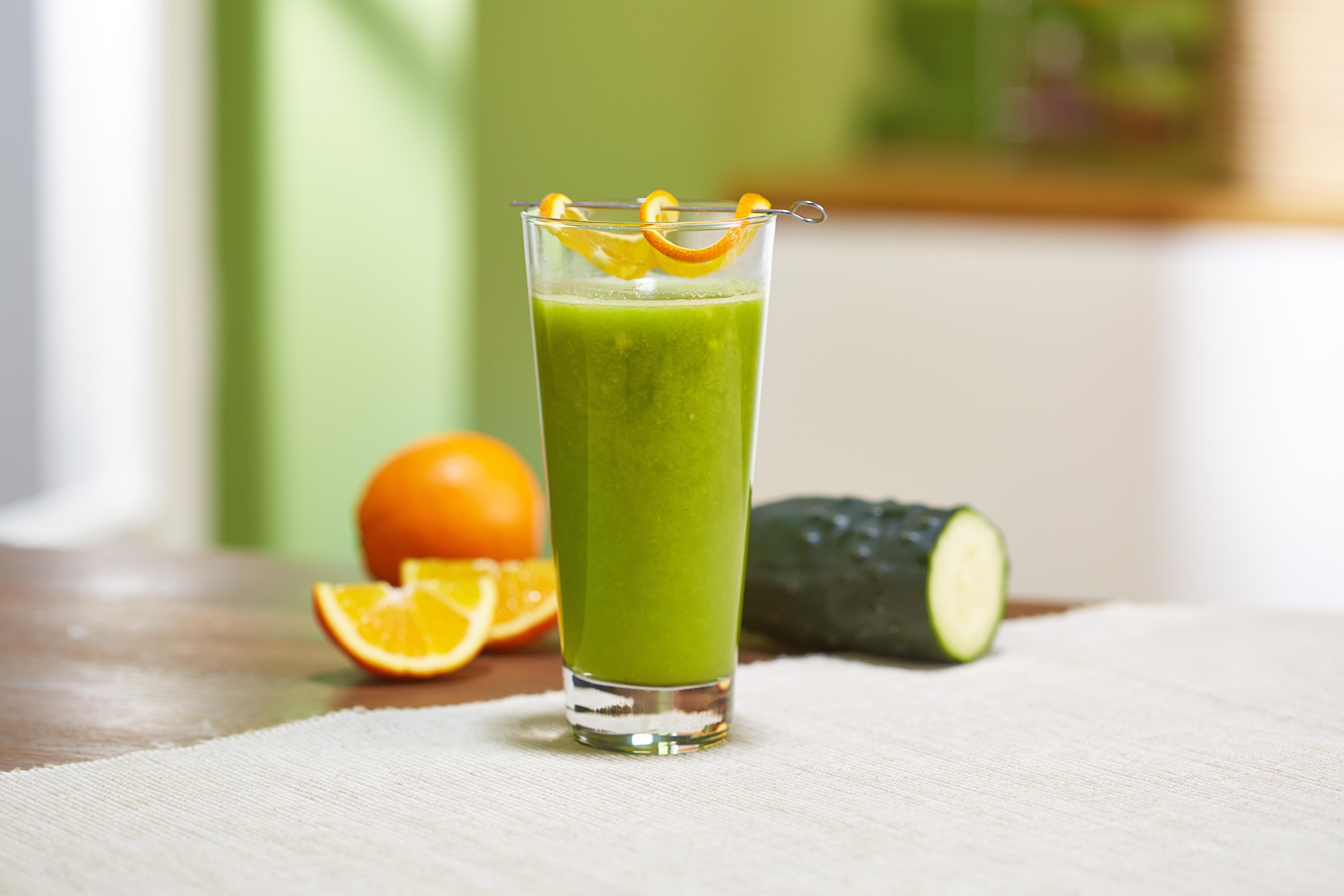 Ein Glas grüner Saft mit Orangendekoration, Orangen und eine Zucchini auf einem Tisch.