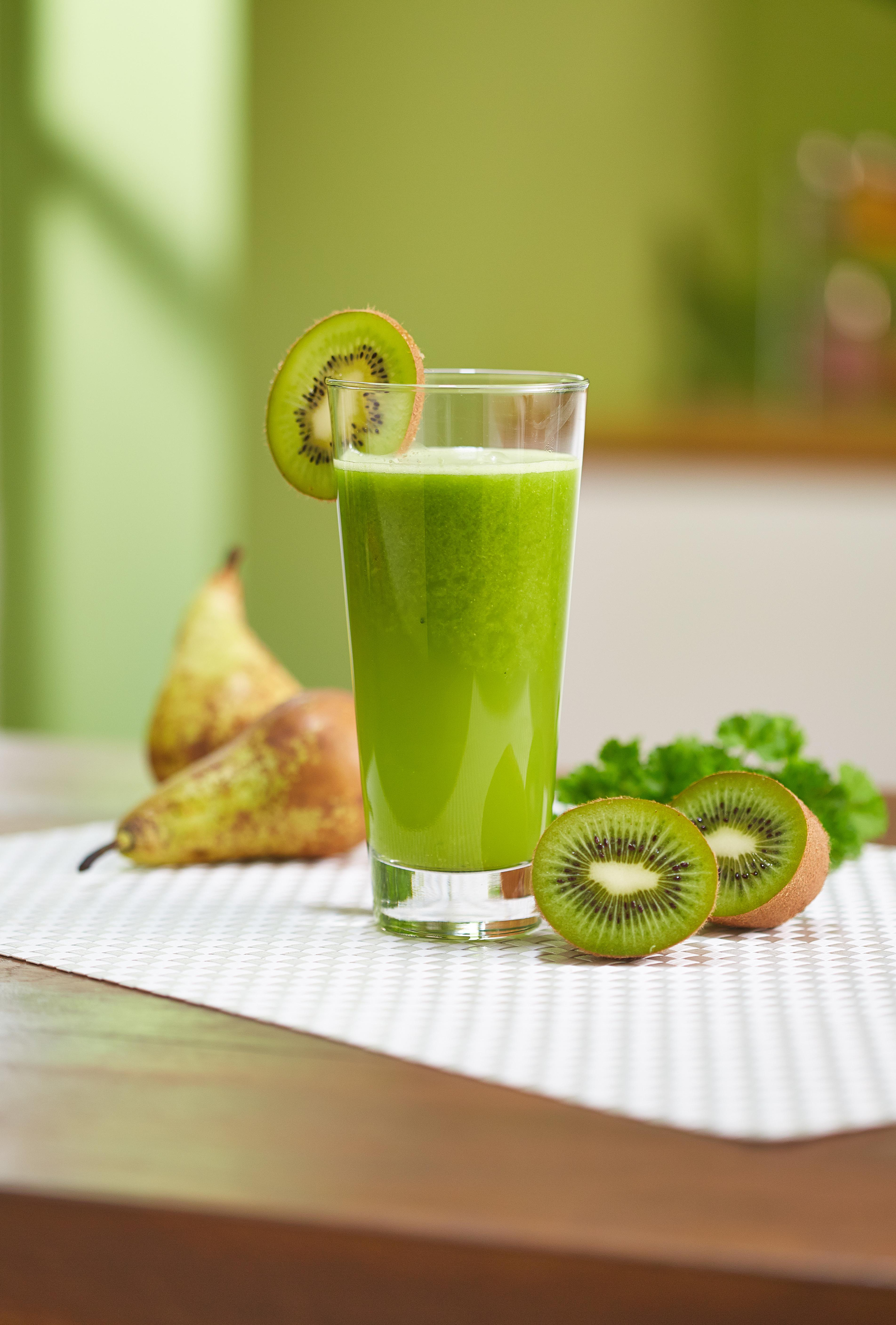 Grüner Smoothie im Glas mit Kiwischeibe, Birnen und Kiwihälften. Weiß gemusterte Matte auf einem Holztisch.