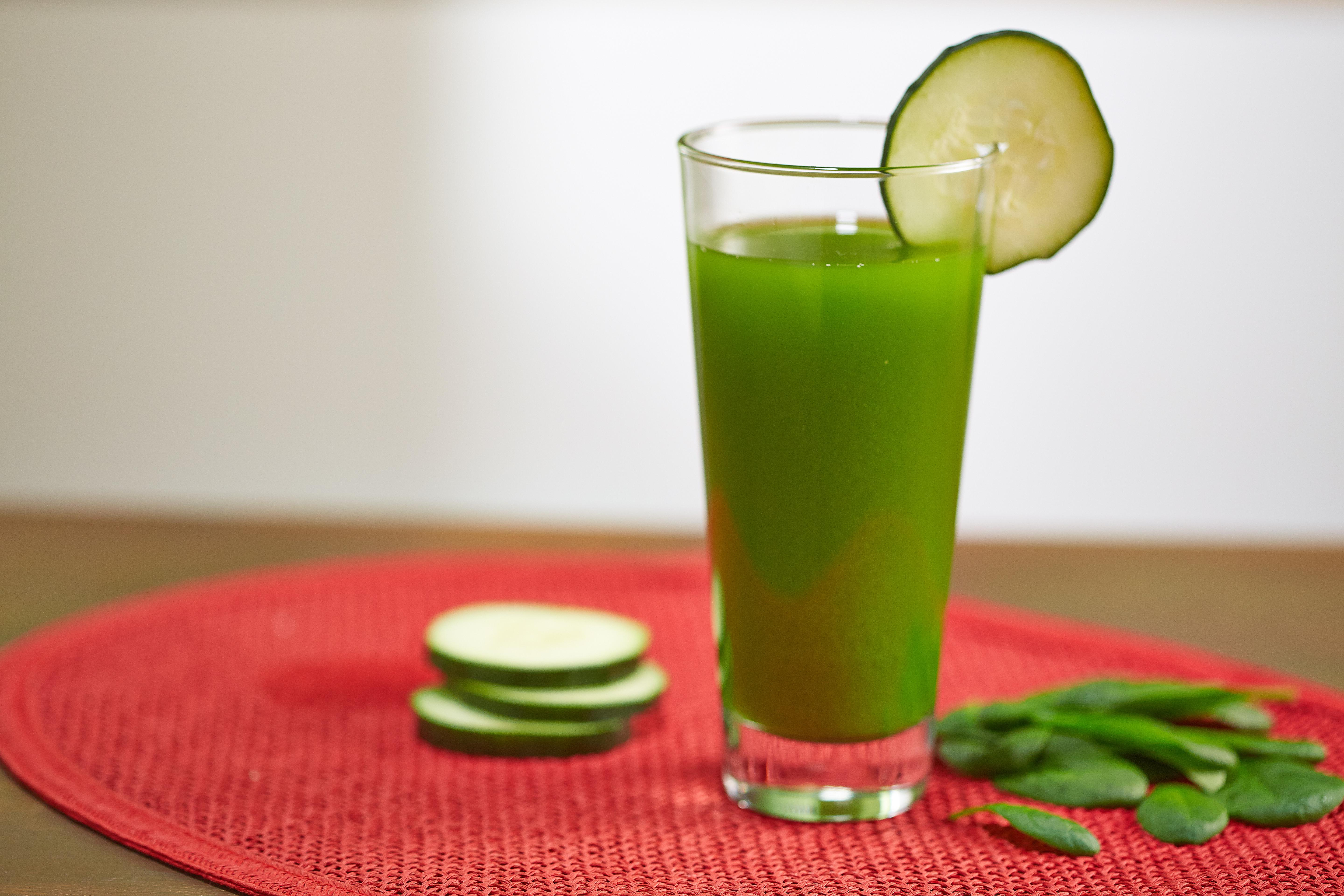 Ein Glas grüner Saft mit Gurkenscheibe, Gurkenscheiben und Spinatblättern auf einer roten Matte.