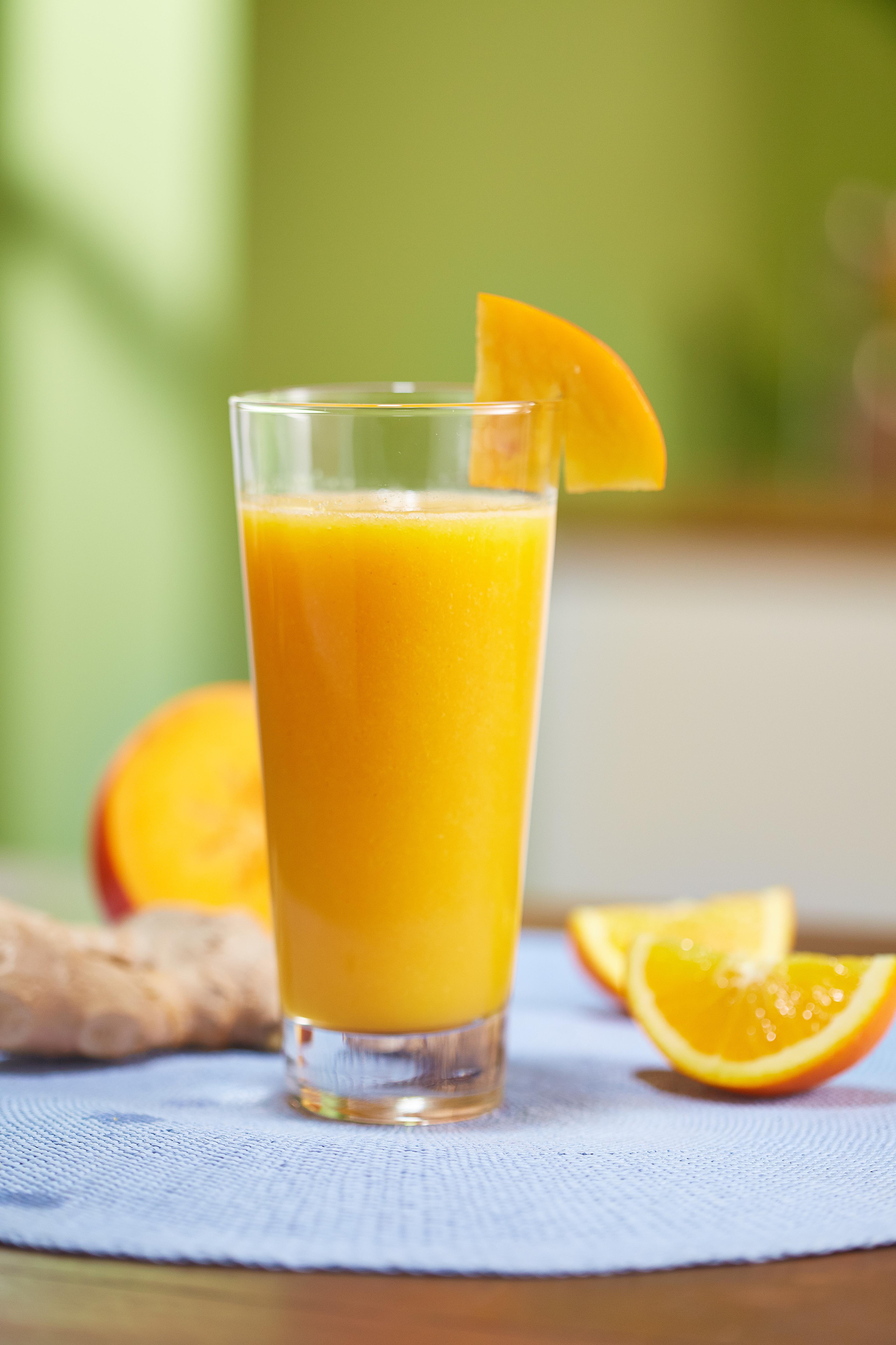 Ein Glas Orangensaft mit einer Orangenscheibe. Orangenstücke und eine Ingwerwurzel liegen auf einem blauen Tuch.