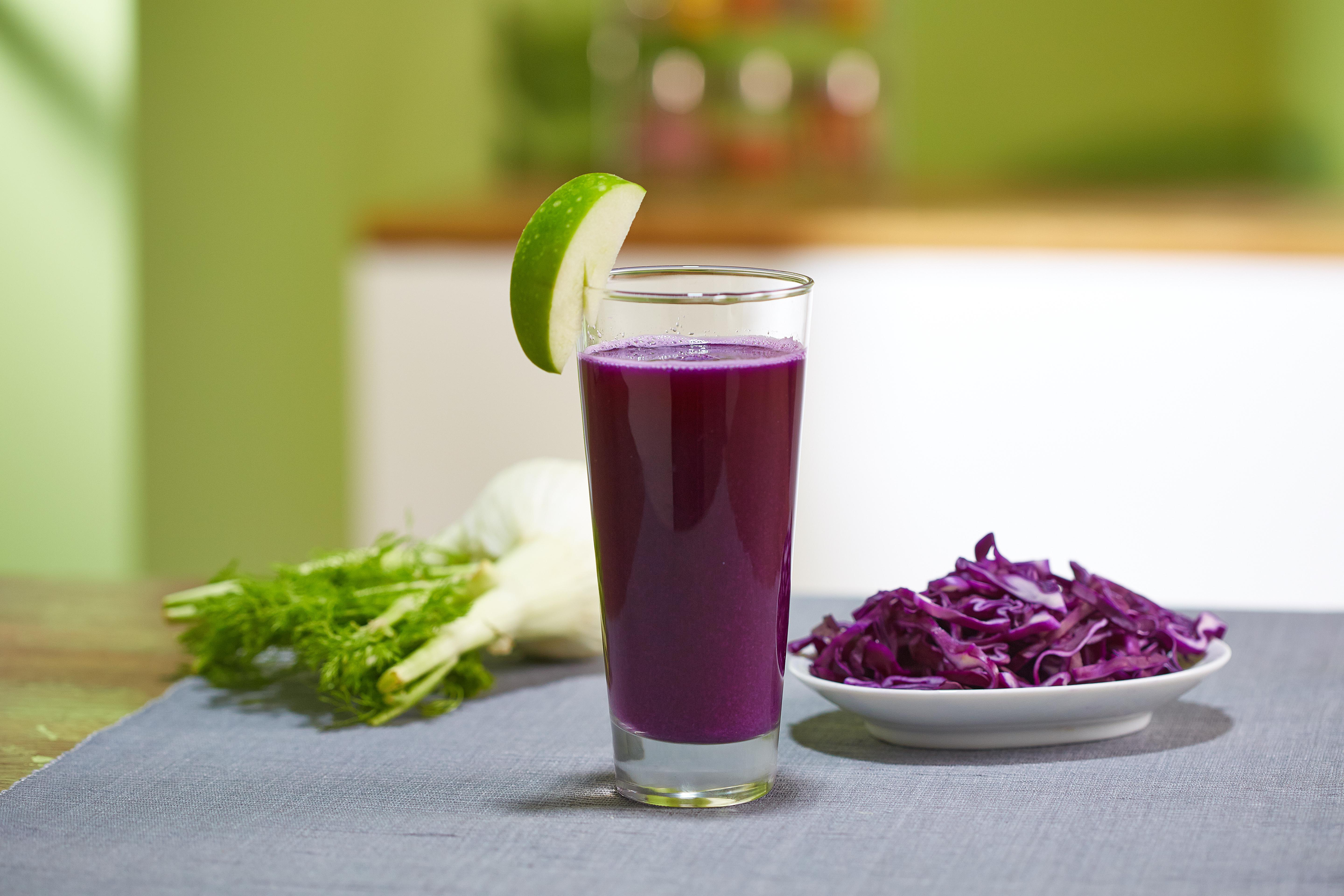 Ein Glas lila Saft mit Apfelscheibe. Rotkohl und Fenchel liegen auf dem Tisch.