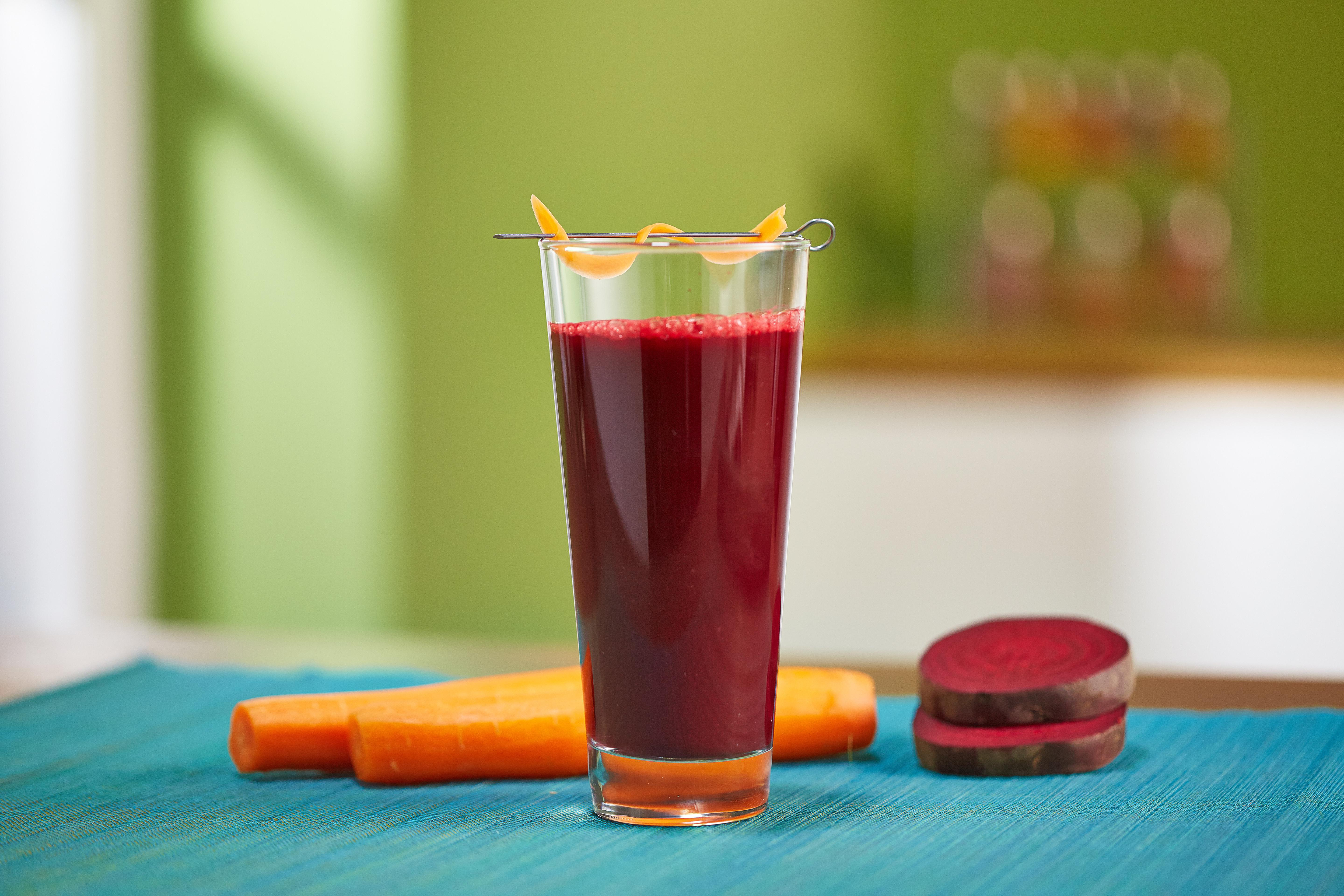 Ein Glas roter Saft mit Karotten-Garnitur, Karotten und Rote-Bete-Scheiben auf einer blauen Matte.