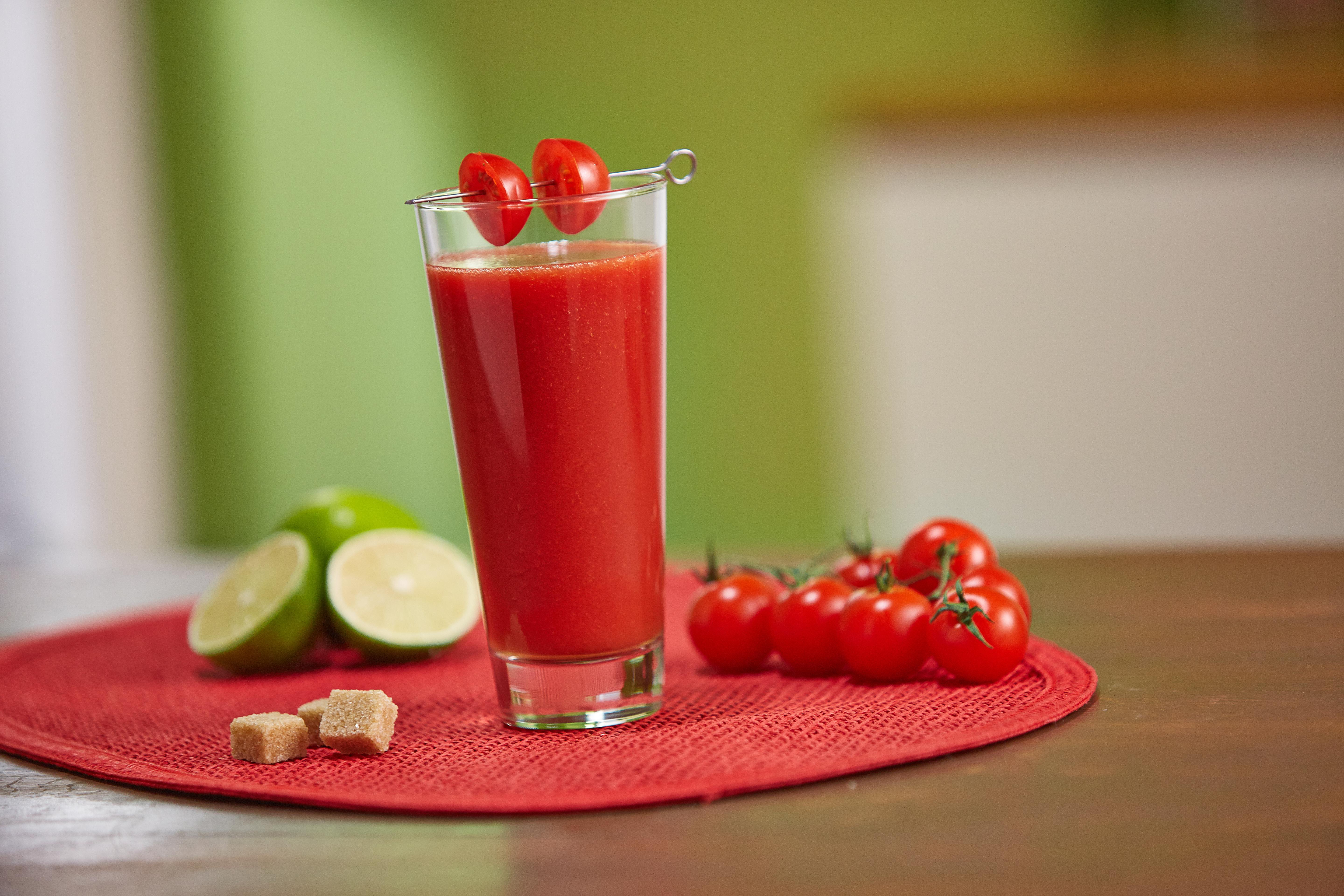 Ein Glas Tomatensaft mit Tomaten oben drauf, Limetten und Würfelzucker auf einer roten Matte. Tomaten sind auch auf der Matte.