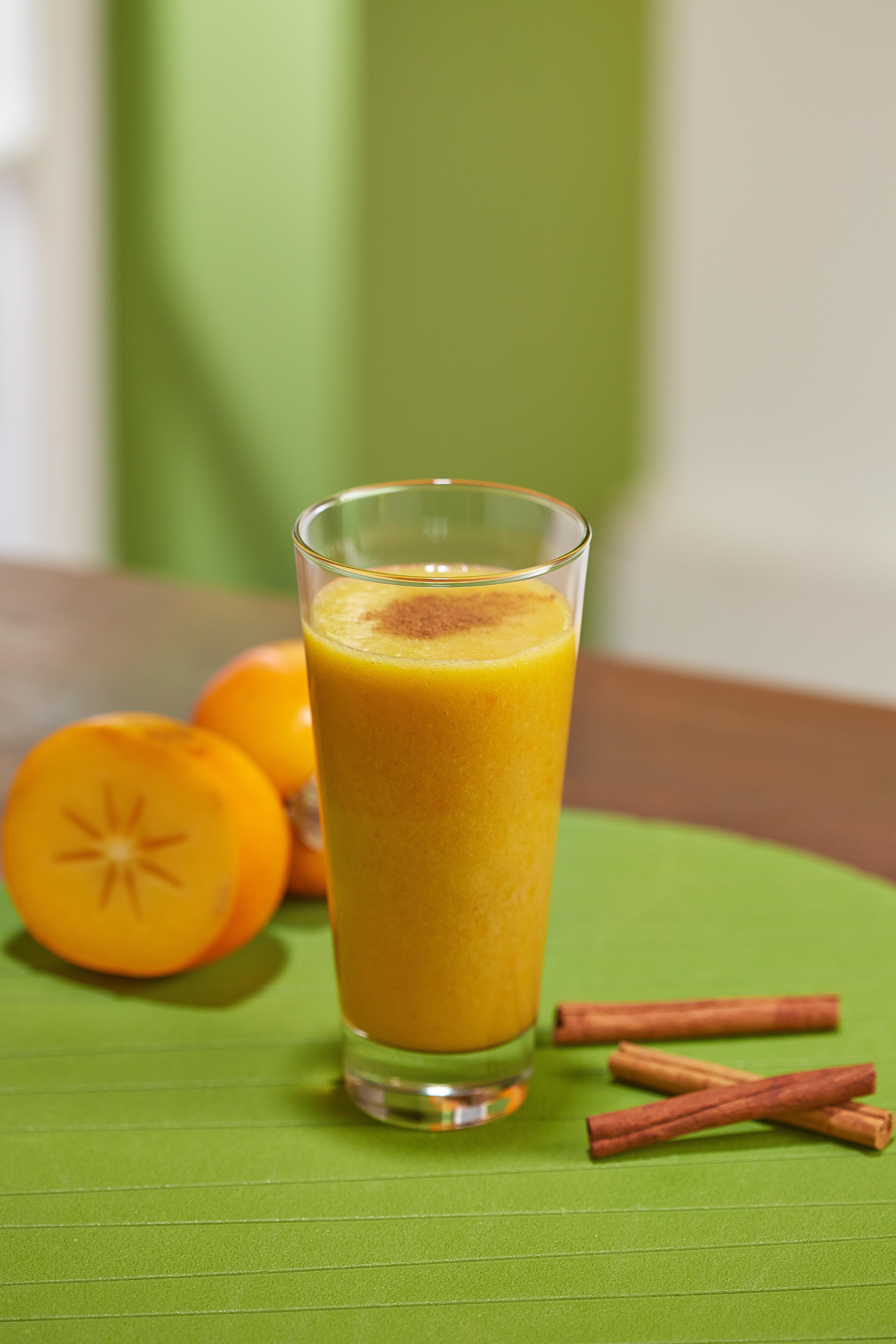 Ein Glas Orangensmoothie mit Zimt, Kaki und Zimtstangen auf einer grünen Oberfläche.