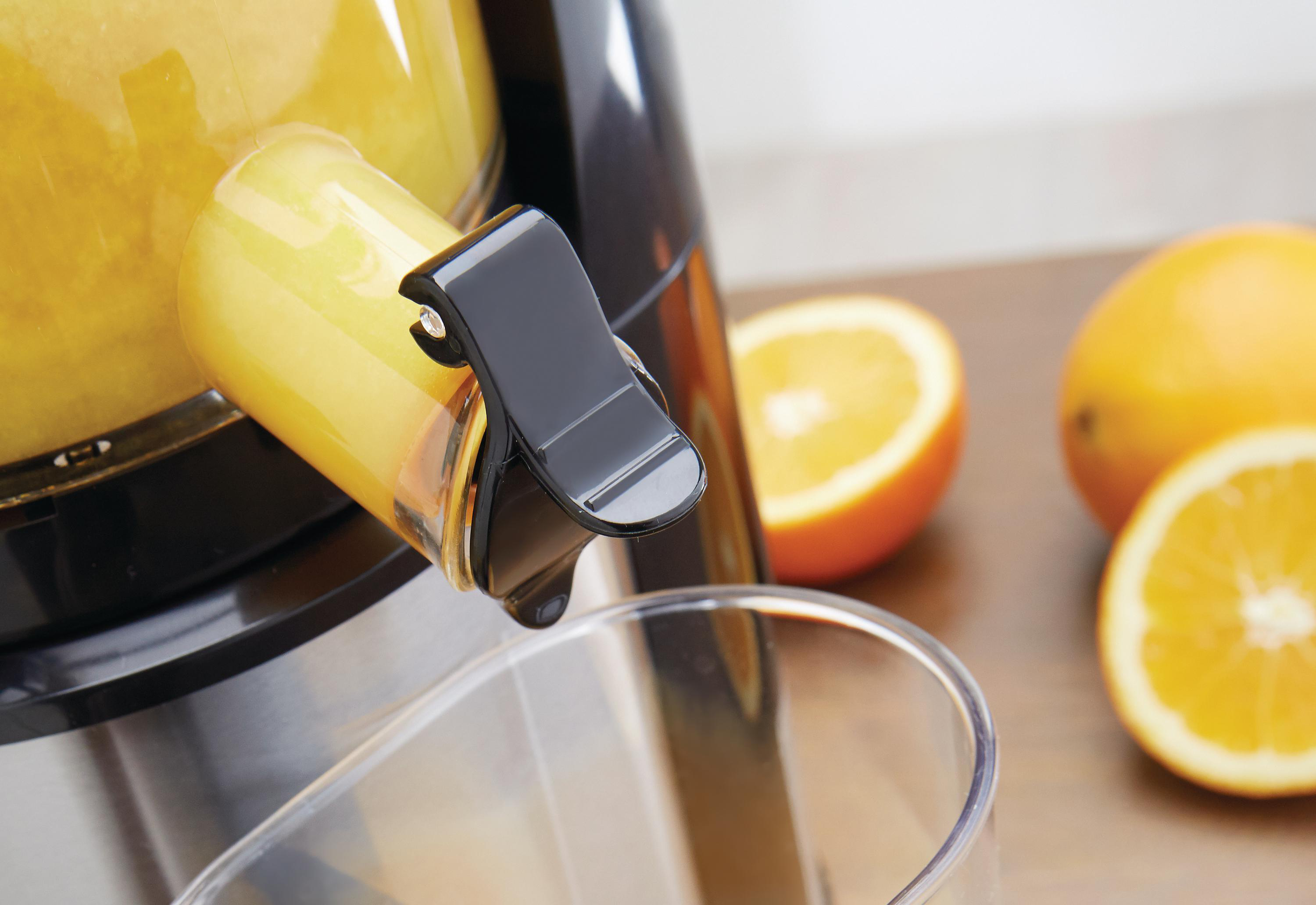Entsafter gießt Saft in ein Glas. Orangen liegen in der Nähe. Der Entsafter ist schwarz und gelb. Glas ist klar.