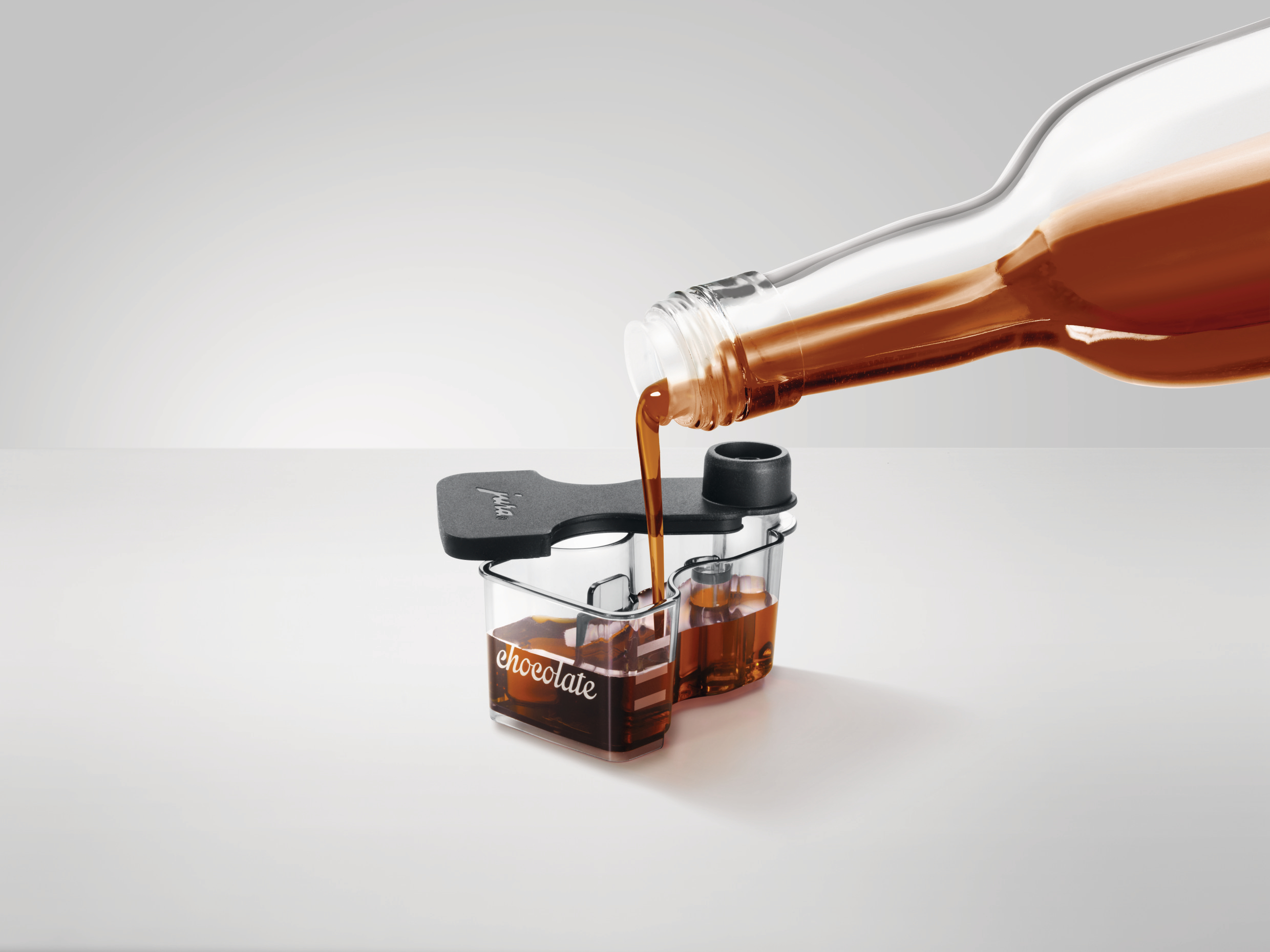 Eine Flasche gießt Schokoladenflüssigkeit in einen Behälter. Der Behälter hat zwei Fächer, eines mit der Aufschrift 'Schokolade'.