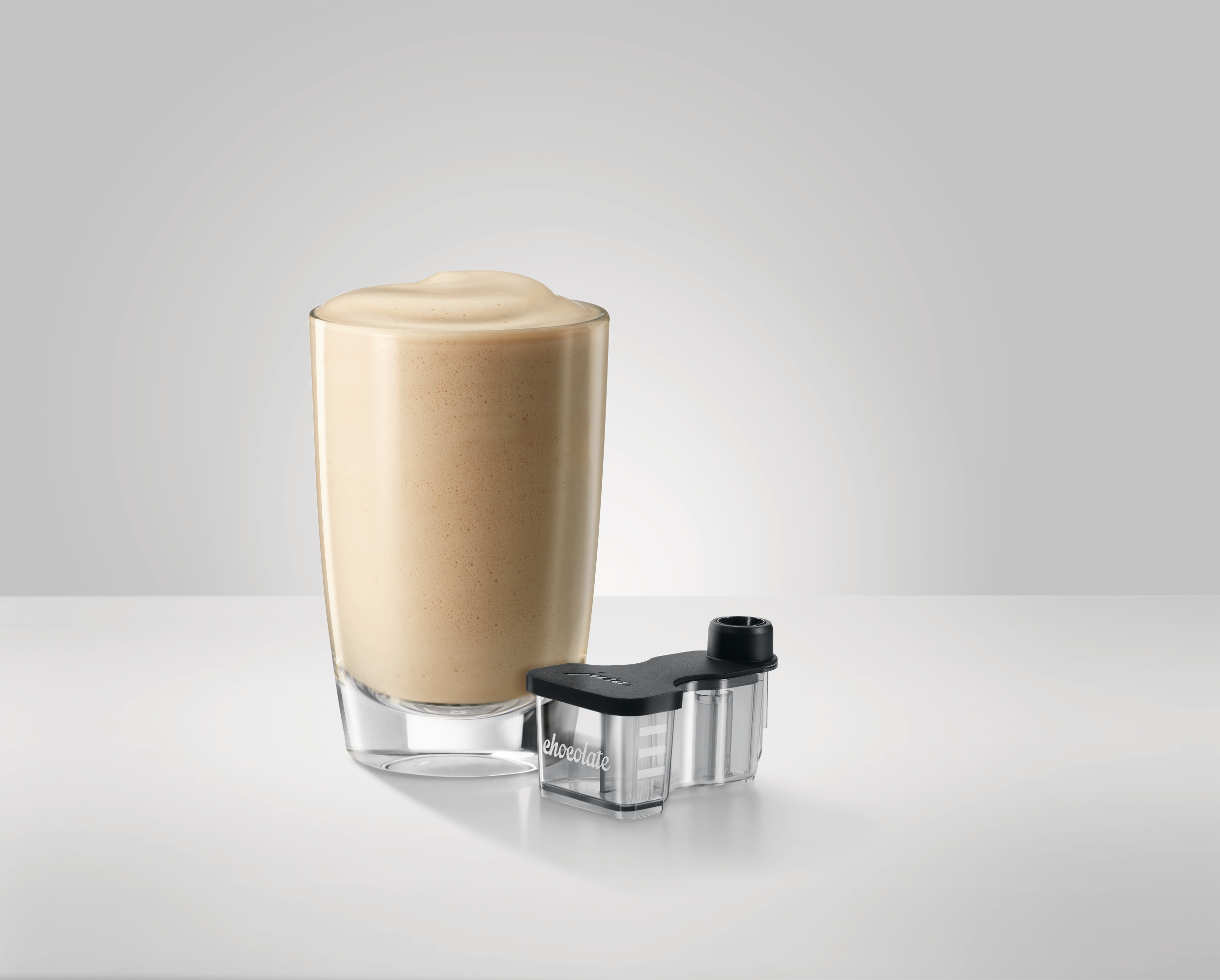 Ein Glas mit braunem Getränk und Schaum, neben einem schwarzen und transparenten Zubehör mit der Aufschrift 'Schokolade'.