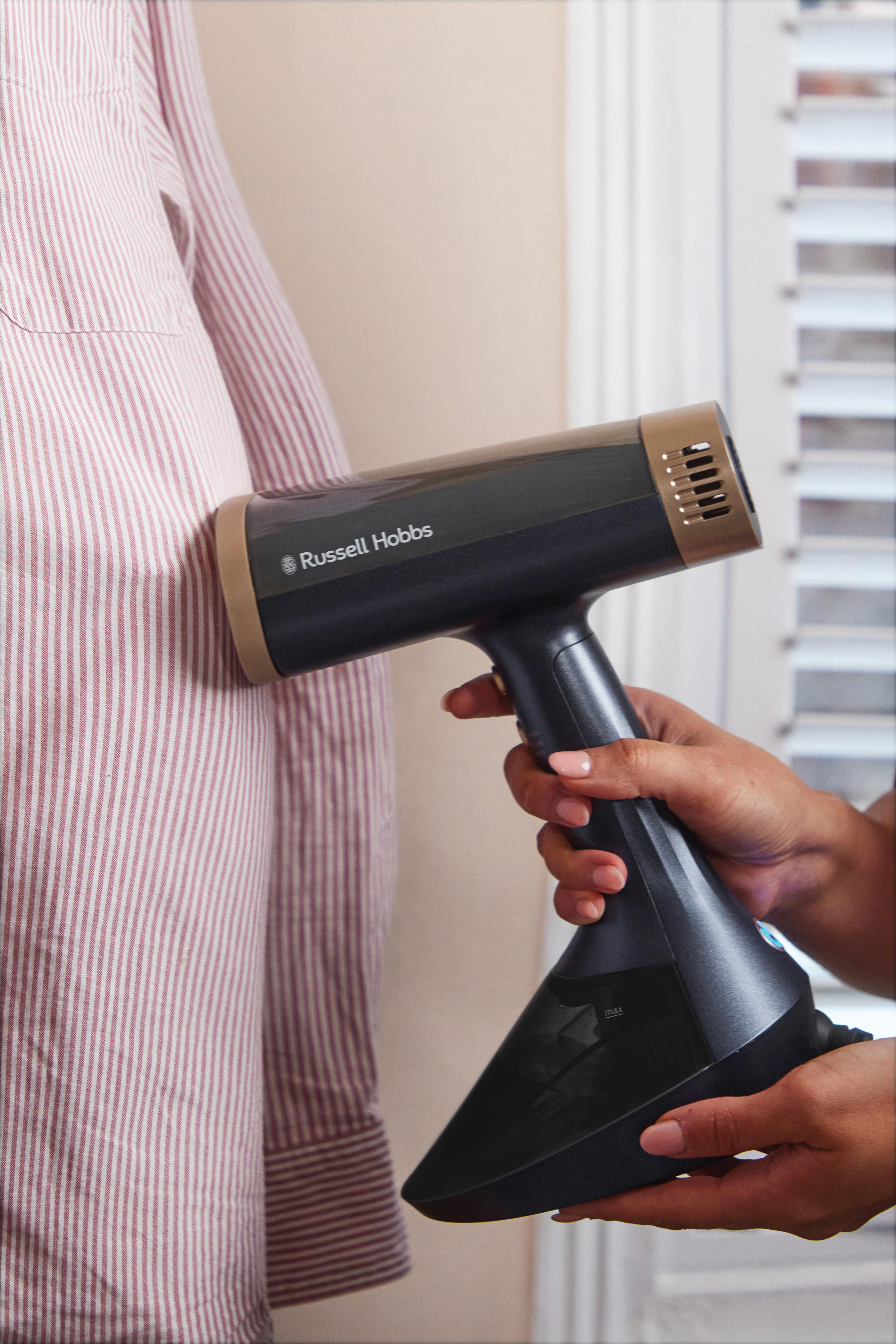 Eine Person bügelt ein gestreiftes Hemd mit einem Russell Hobbs Dampfgarer. Der Hintergrund ist eine Wand und ein Fenster mit Jalousien.