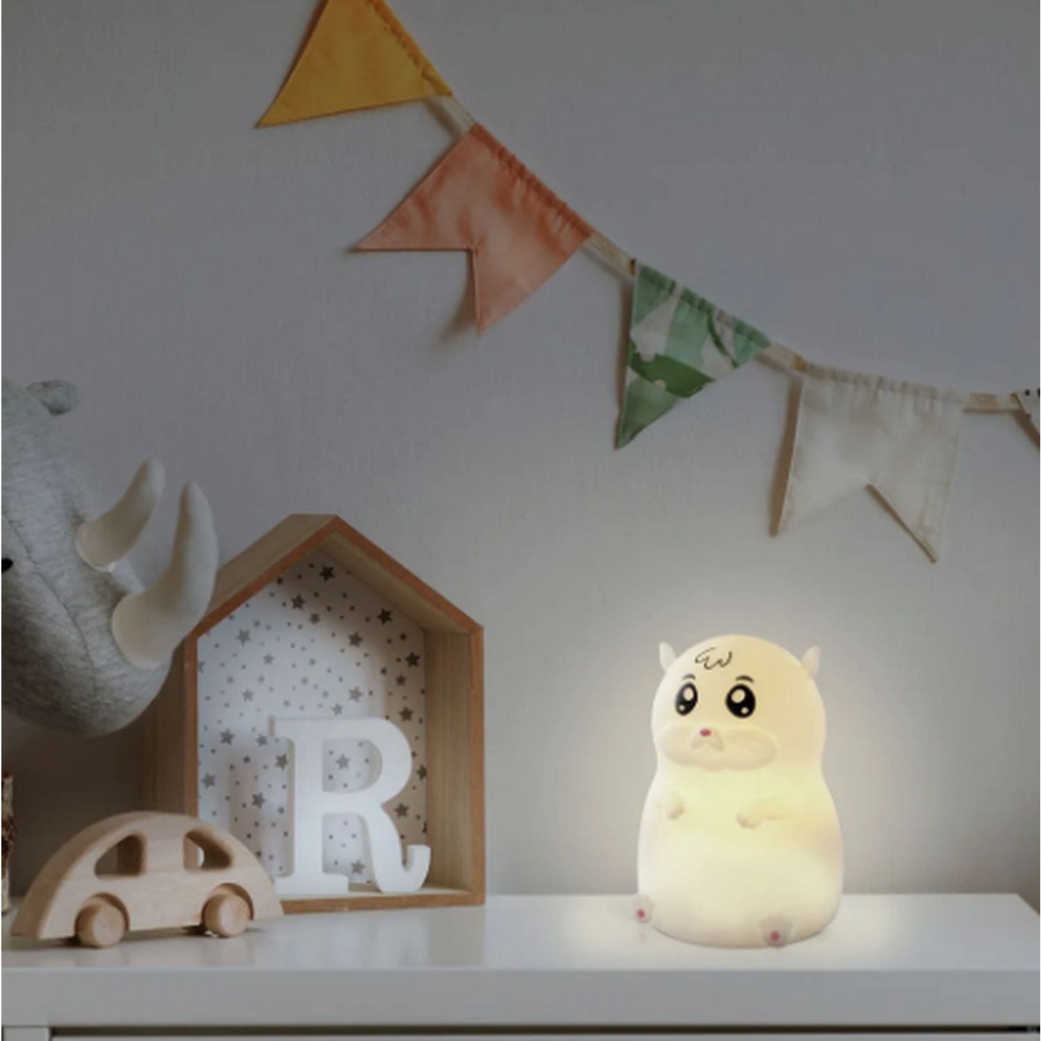 A hamster-shaped night light sits on a white surface. A wooden car, a letter 'R', and a house-shaped shelf are also visible. Colorful flags hang above.
