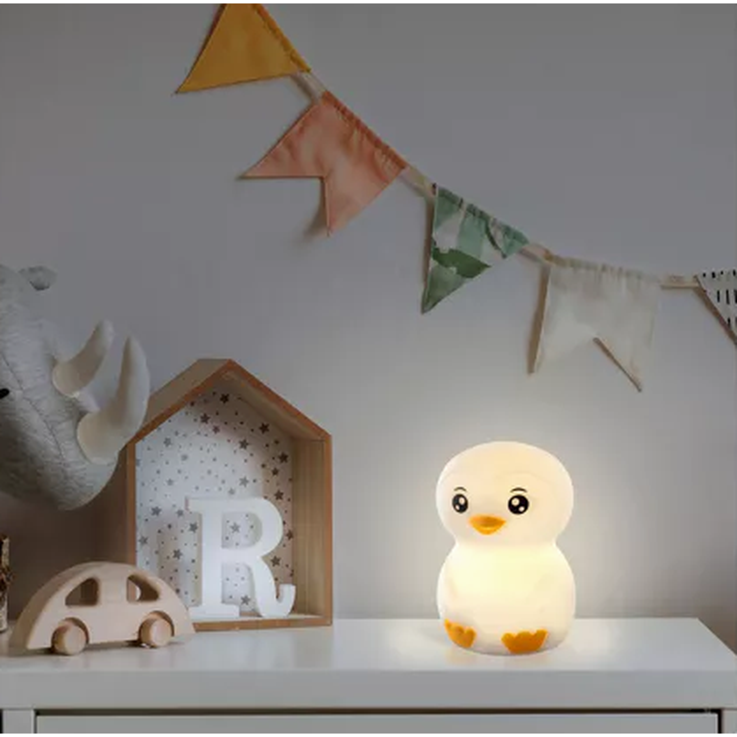 A penguin-shaped night light is on a white surface. A wooden car and house are also on the surface. Colorful flags hang above.