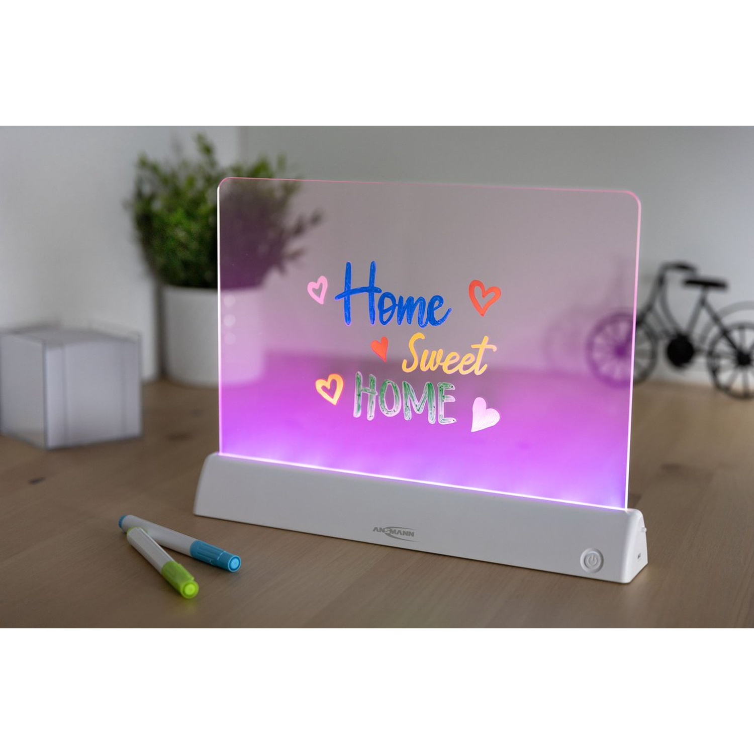 Een verlicht bord met 'Home Sweet Home' met kleurrijke harten, pennen en een plant. Een fietssilhouet is zichtbaar op de achtergrond.