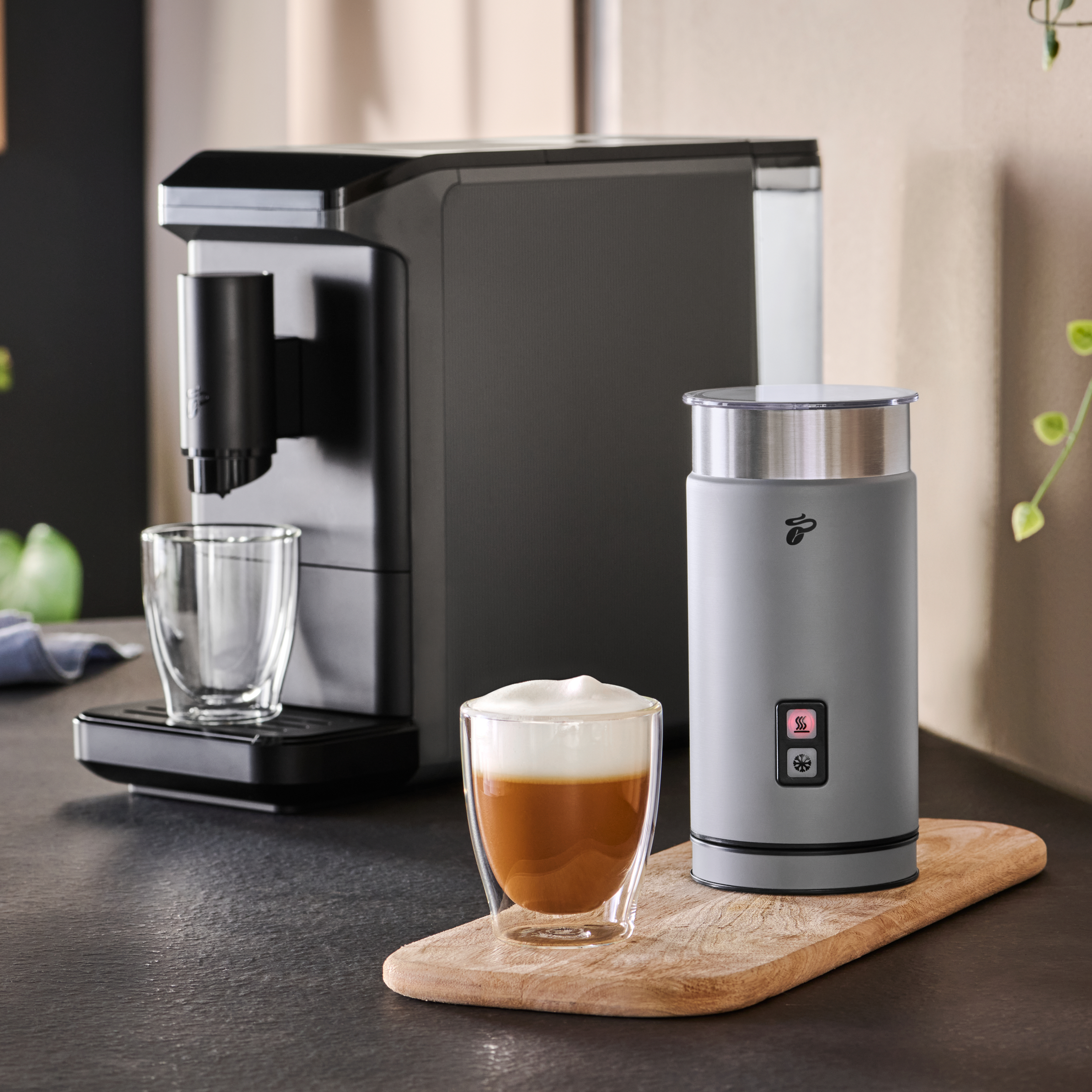 Kaffeemaschine und Milchaufschäumer auf einem Holzbrett. Ein Glas Kaffee mit Milchschaum steht auf dem Brett.