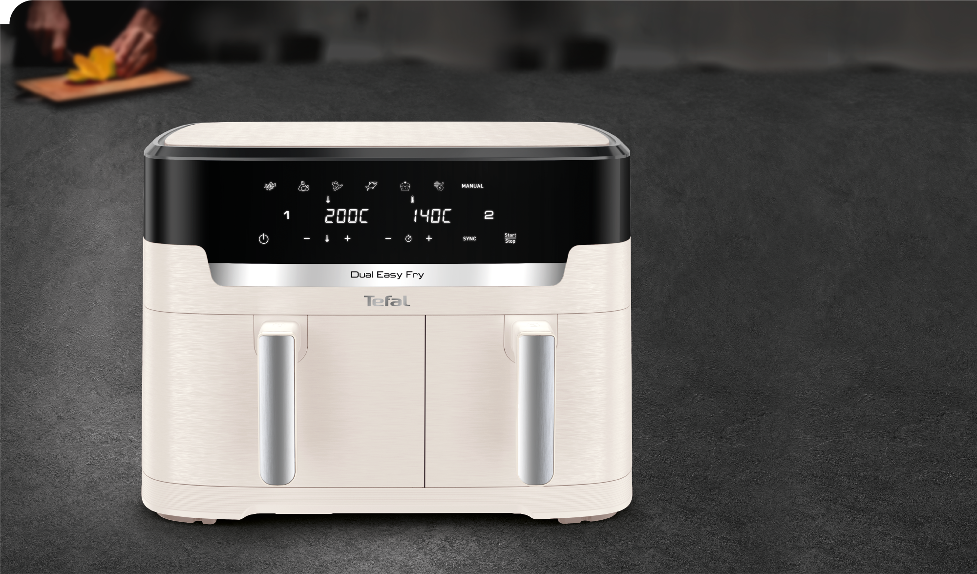 Krémszínű Tefal Dual Easy Fry fritőz sötét felületen. Egy személy gyümölcsöt vág az elmosódott háttérben.