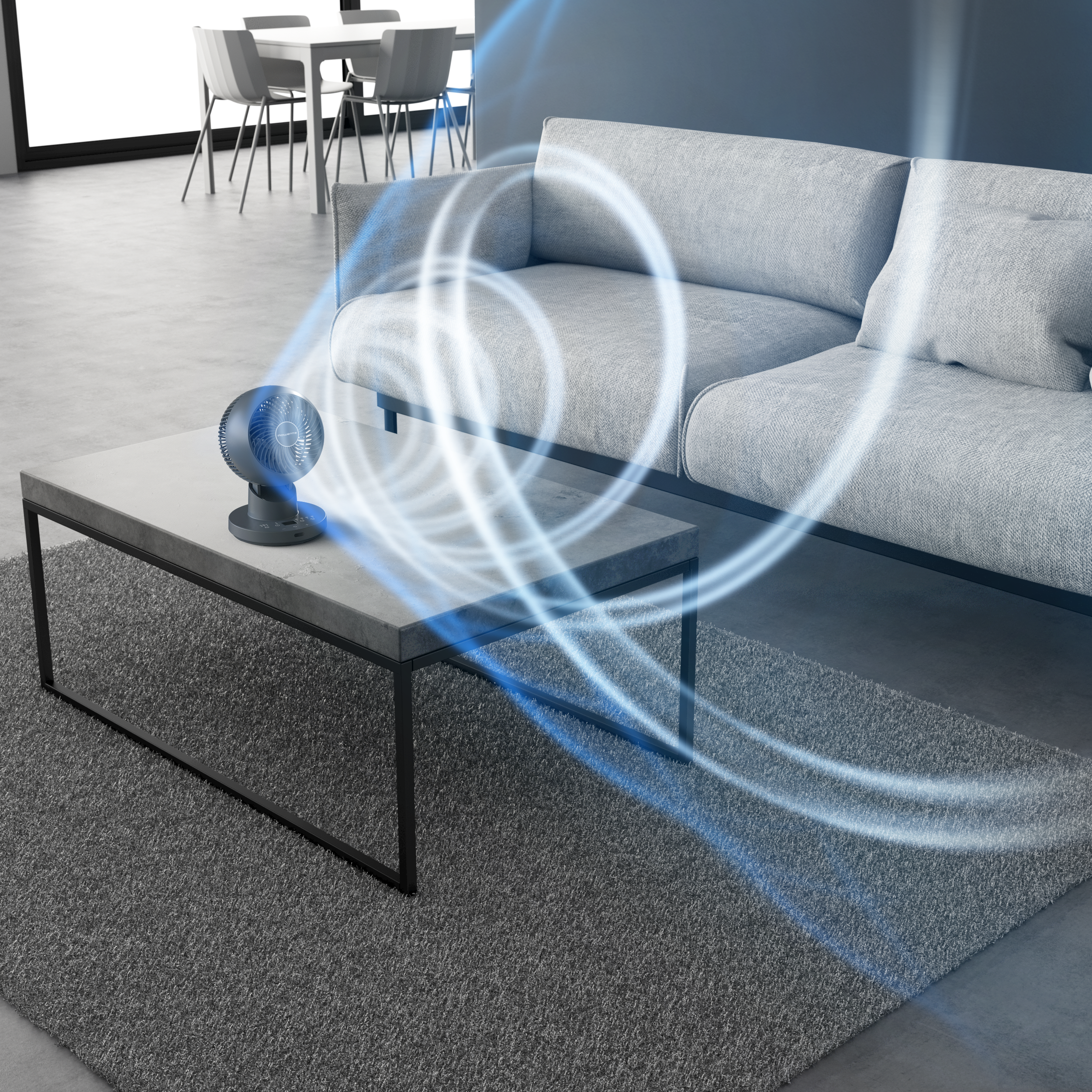 Egy szürke ventilátor egy asztalon légáramlási vonalakkal. Jobbra egy szürke kanapé. Hátul egy étkezőasztal és székek.