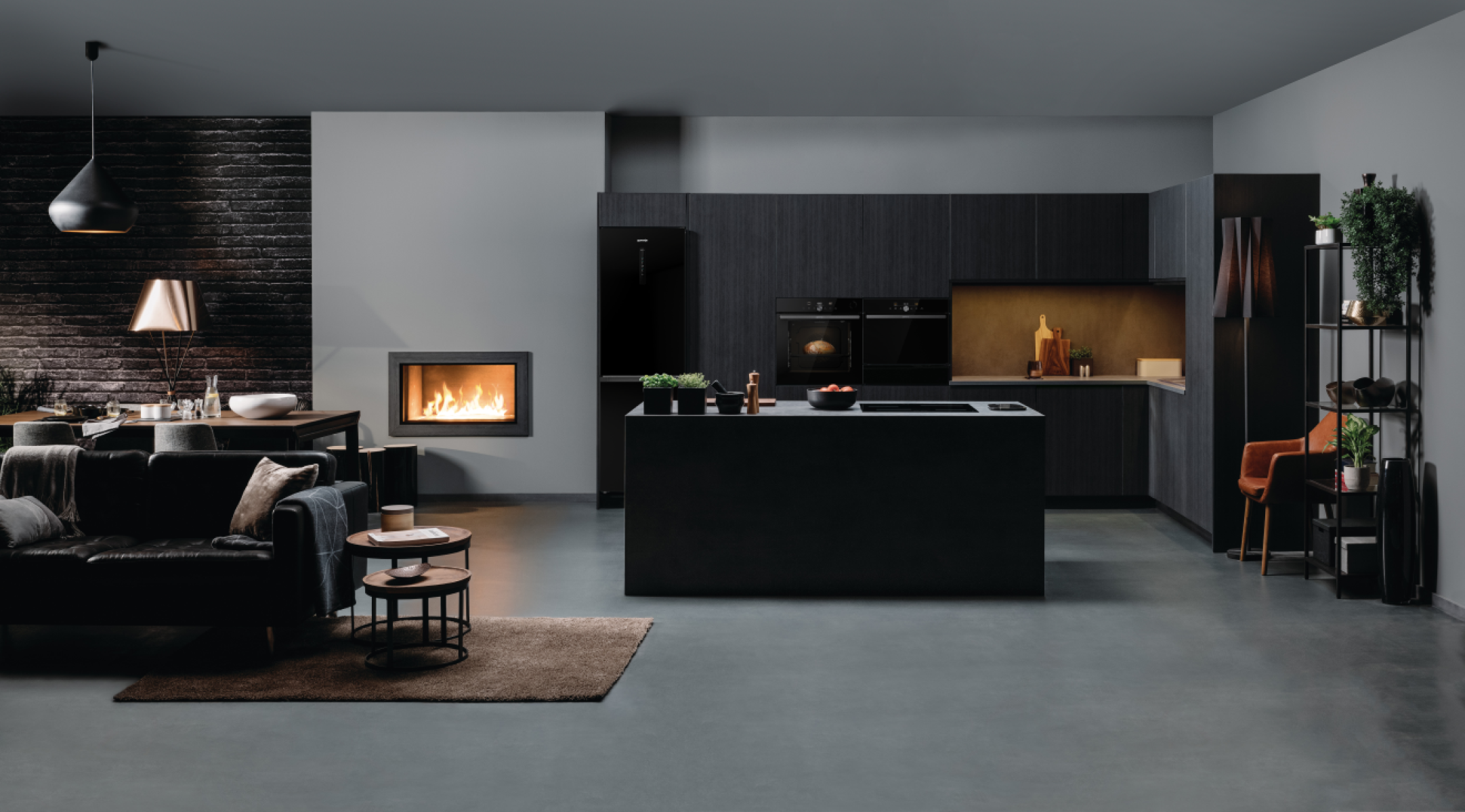Nowoczesna kuchnia z ciemnymi szafkami, wyspą i kominkiem. Zawiera czarną lodówkę i piekarnik. Salon z sofą i stołem.