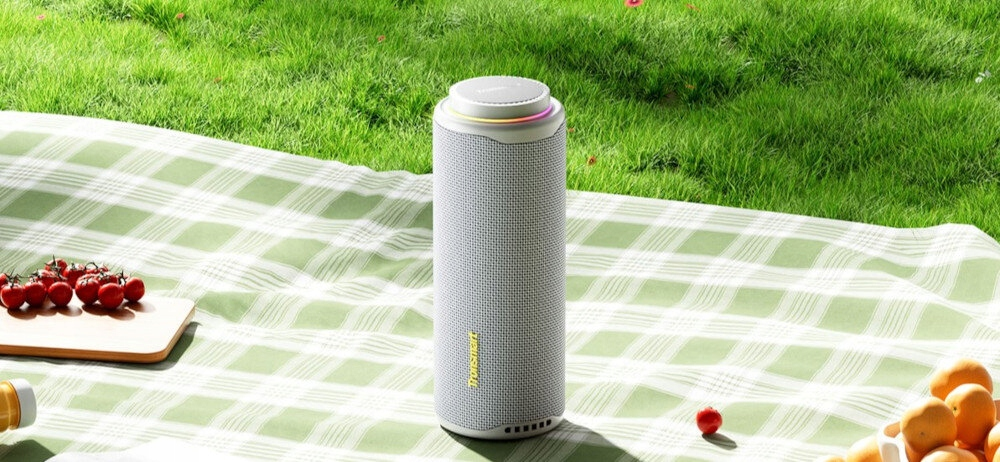 Szary głośnik stoi na kocu piknikowym z pomidorami i pomarańczami. Zielona trawa w tle.