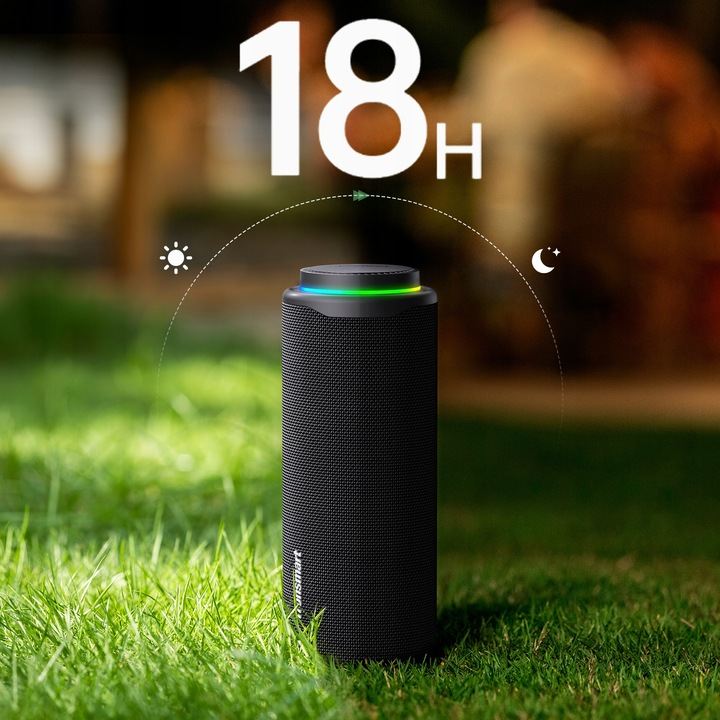 Czarny głośnik z kolorowym wierzchem stoi na zielonej trawie. Tło jest rozmyte, z tekstem '18h' nad głośnikiem.