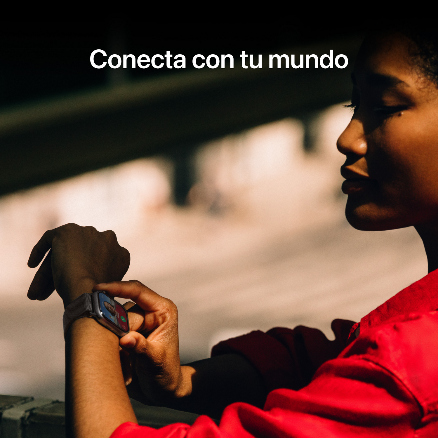 Una mujer con camisa roja mira su Apple Watch, que muestra una videollamada. El fondo está borroso, con texto 'Conecta con tu mundo'.