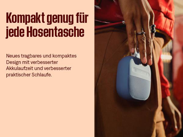 Eine Person hält einen kleinen blauen Lautsprecher. Der Lautsprecher befindet sich vor einer Person, die eine rote Jacke und braune Hose trägt.