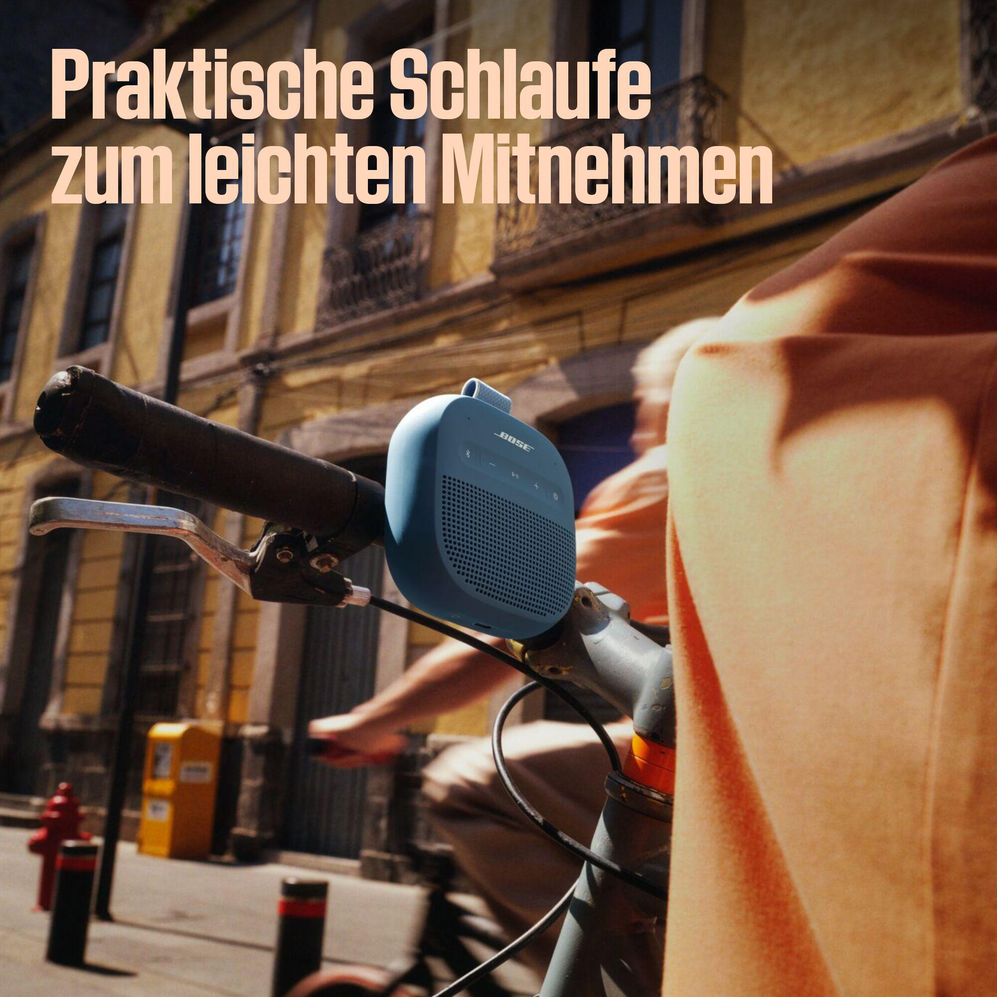 Ein blauer Bose-Lautsprecher ist an einem Fahrradlenker befestigt. Der Hintergrund zeigt eine Straßenszene mit Gebäuden und einem Radfahrer.