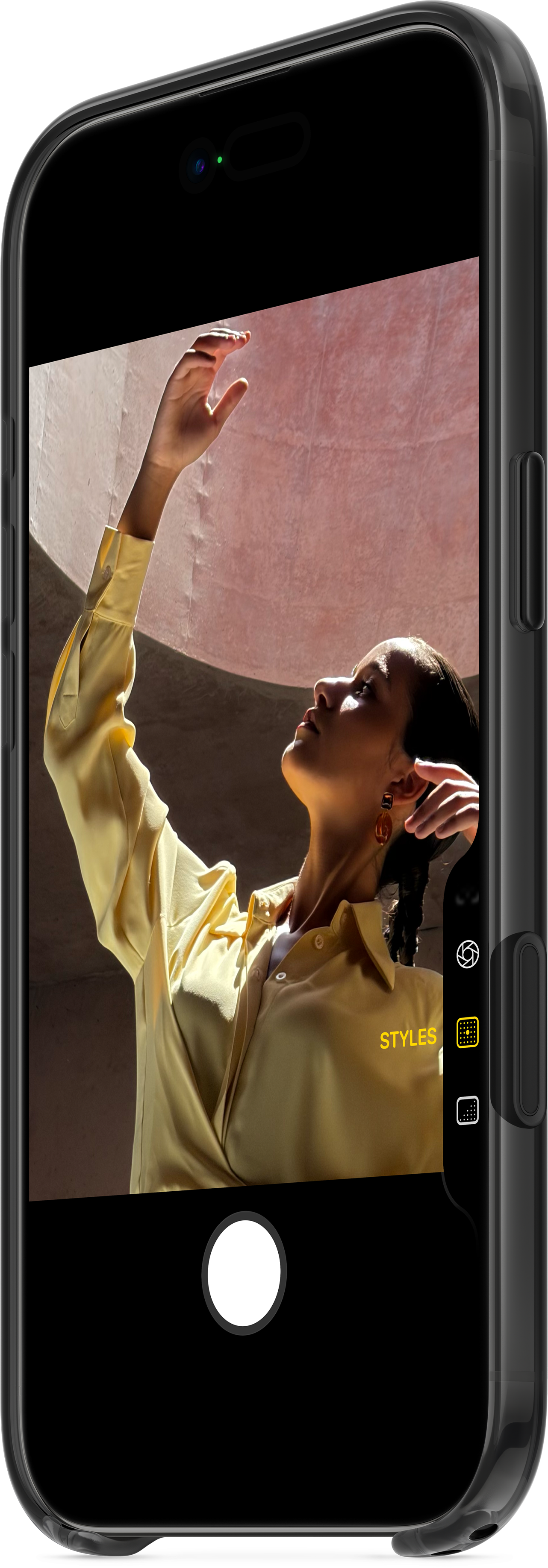 Frau im gelben Hemd, Hand erhoben, auf einem Telefonbildschirm. Rosa Hintergrund. Telefon ist schwarz mit Kamerasteuerung.