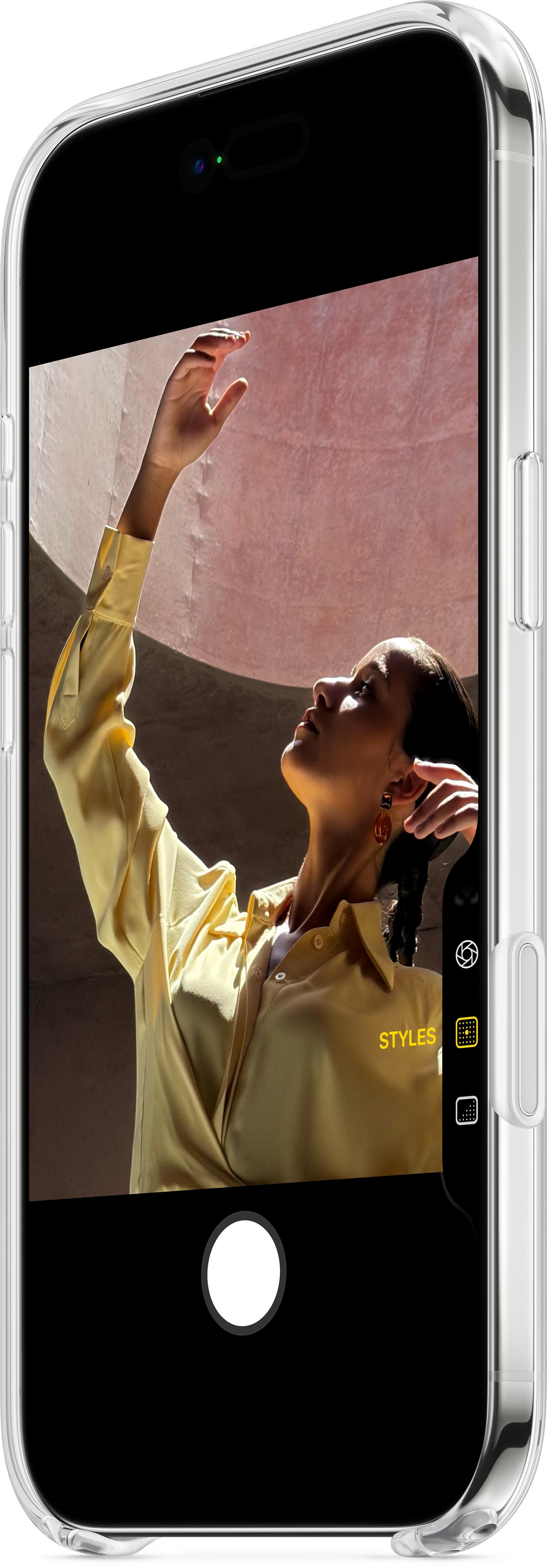Femme en chemise jaune pose, levant la main. Le téléphone affiche l'image. Fond rose.