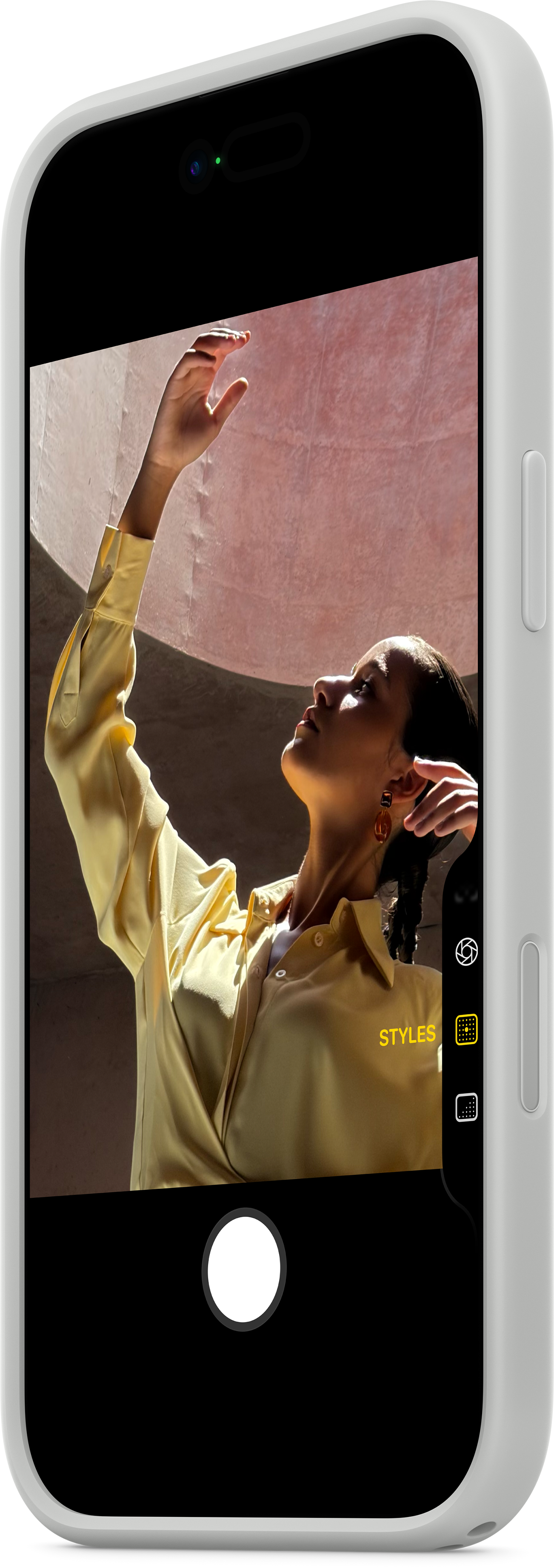 Frau im gelben Hemd hebt den Arm und posiert für ein Foto auf einem Telefonbildschirm. Rosa Wand im Hintergrund, Telefon in weißem Gehäuse.