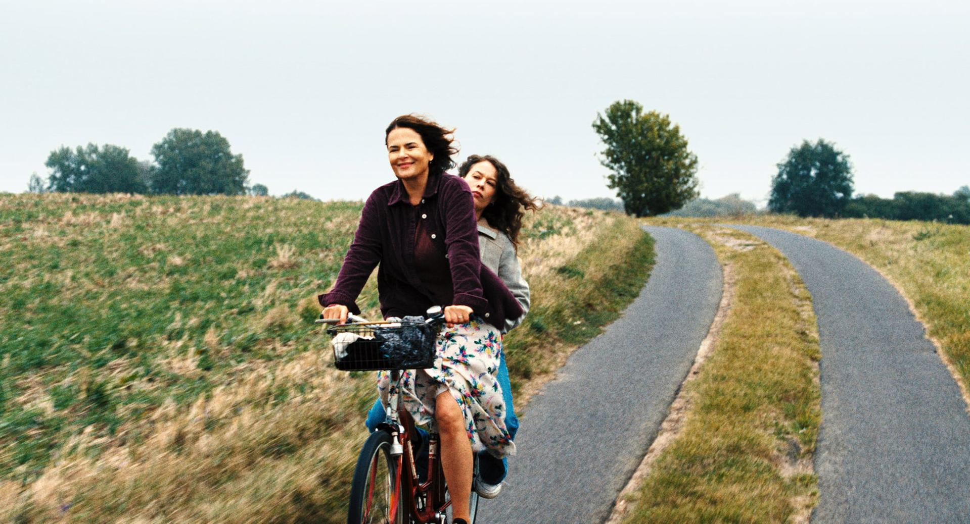 Zwei Frauen fahren mit dem Fahrrad auf einem asphaltierten Weg. Eine lenkt, die andere sitzt hinten. Grüne Felder und Bäume umgeben den Weg.