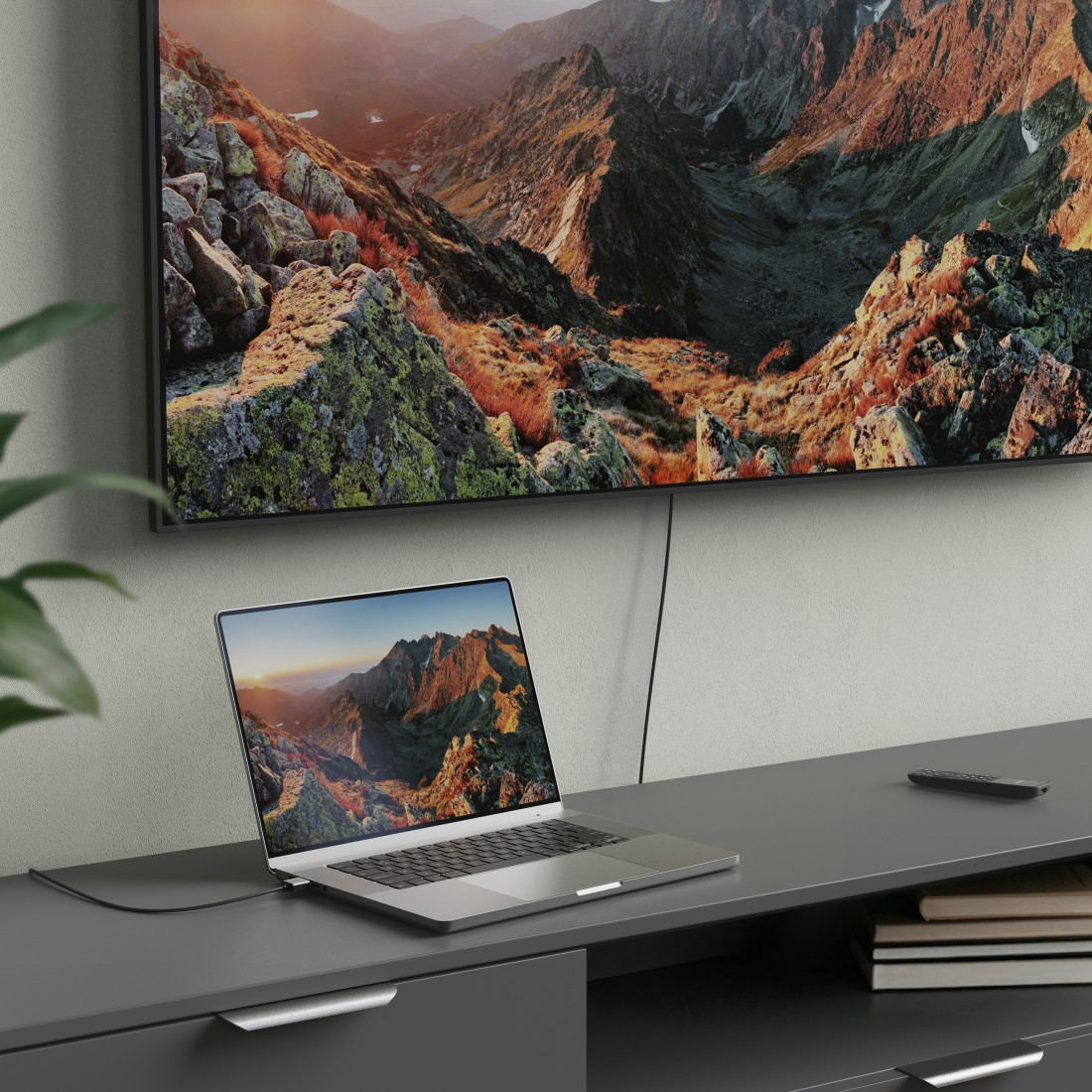 Laptop auf dunklem Schreibtisch. Ein großer Fernseher zeigt eine Berglandschaft. Bücher und eine Fernbedienung sind in der Nähe.