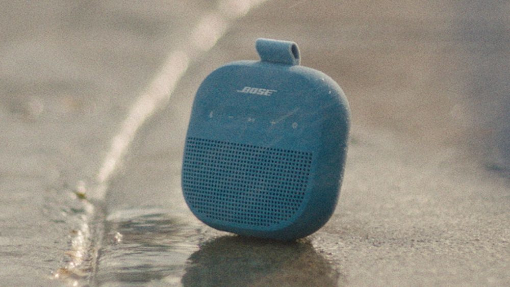 Niebieski głośnik Bose stoi na mokrej powierzchni. Głośnik ma uchwyt i kratkę. Tło jest rozmyte.