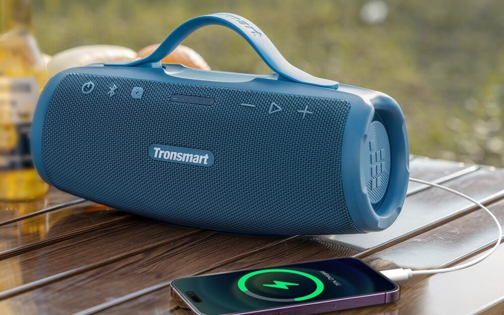 Niebieski głośnik Tronsmart ładujący telefon na drewnianym stole na zewnątrz. Tło rozmyte, butelka i zieleń.