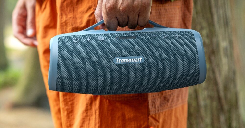 Osoba trzyma niebieski głośnik Tronsmart z uchwytem. Osoba ma na sobie pomarańczowe spodnie. Tło jest rozmyte zielone.