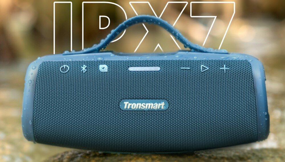 Niebieski głośnik Tronsmart z uchwytem, przyciskami i logo. Krople wody na nim. Tło jest rozmyte.