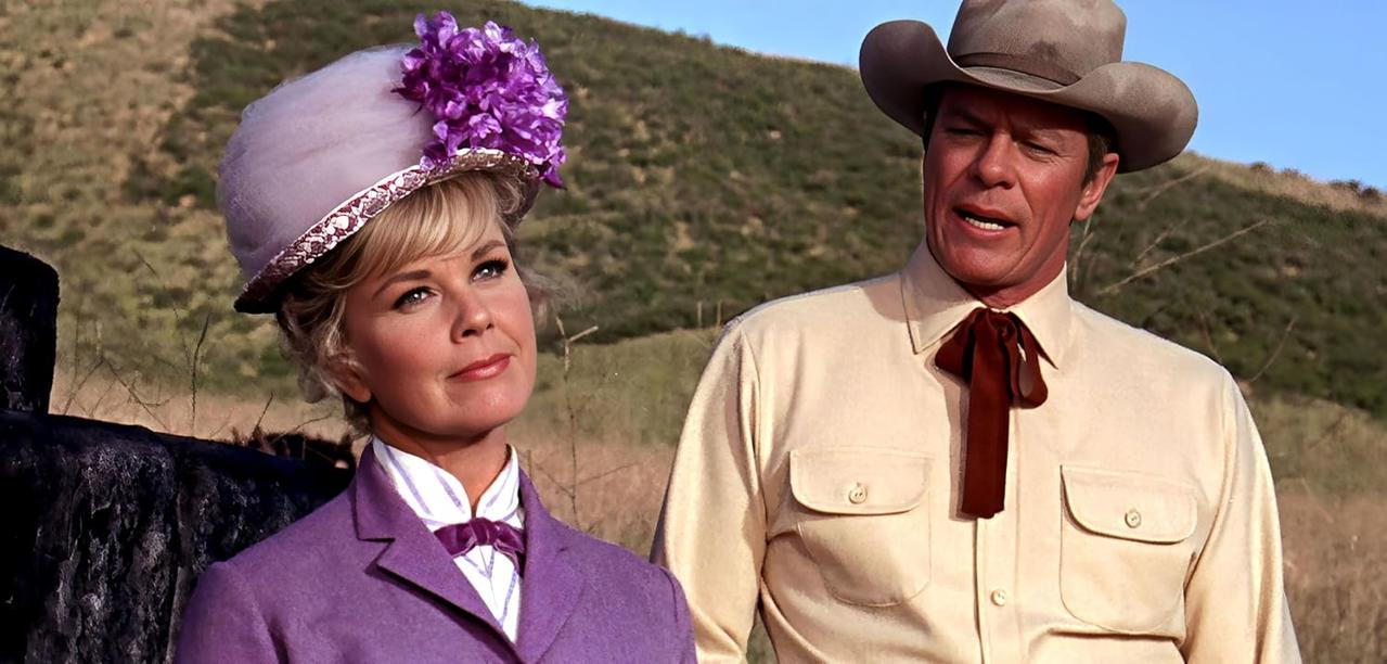 Eine Frau in lila Outfit und Hut mit einem Mann in einem Cowboyhut, stehend auf einem Feld. Im Hintergrund ist ein Hügel und blauer Himmel.