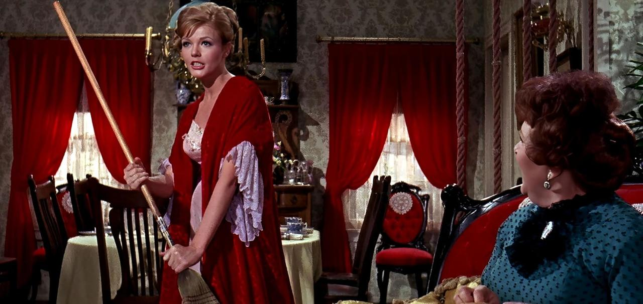 Eine Frau in einem roten Bademantel hält einen Besen und blickt eine andere Frau in einem blauen Kleid an. Rote Vorhänge und Möbel im Hintergrund.