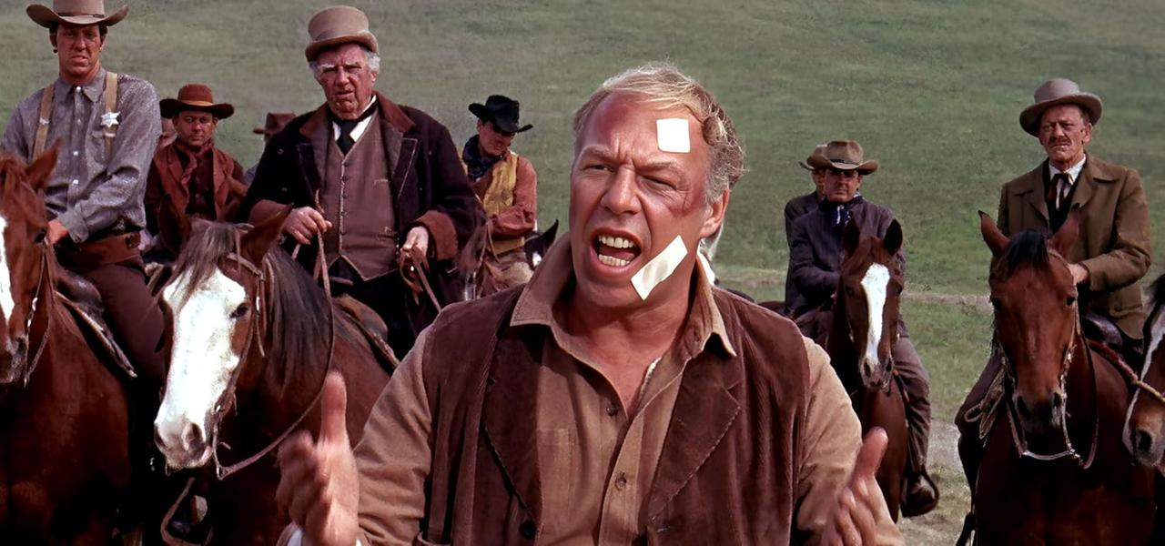Eine Gruppe von Männern auf Pferden, einige tragen Cowboyhüte. Ein Mann vorne hat einen Verband im Gesicht und schreit.